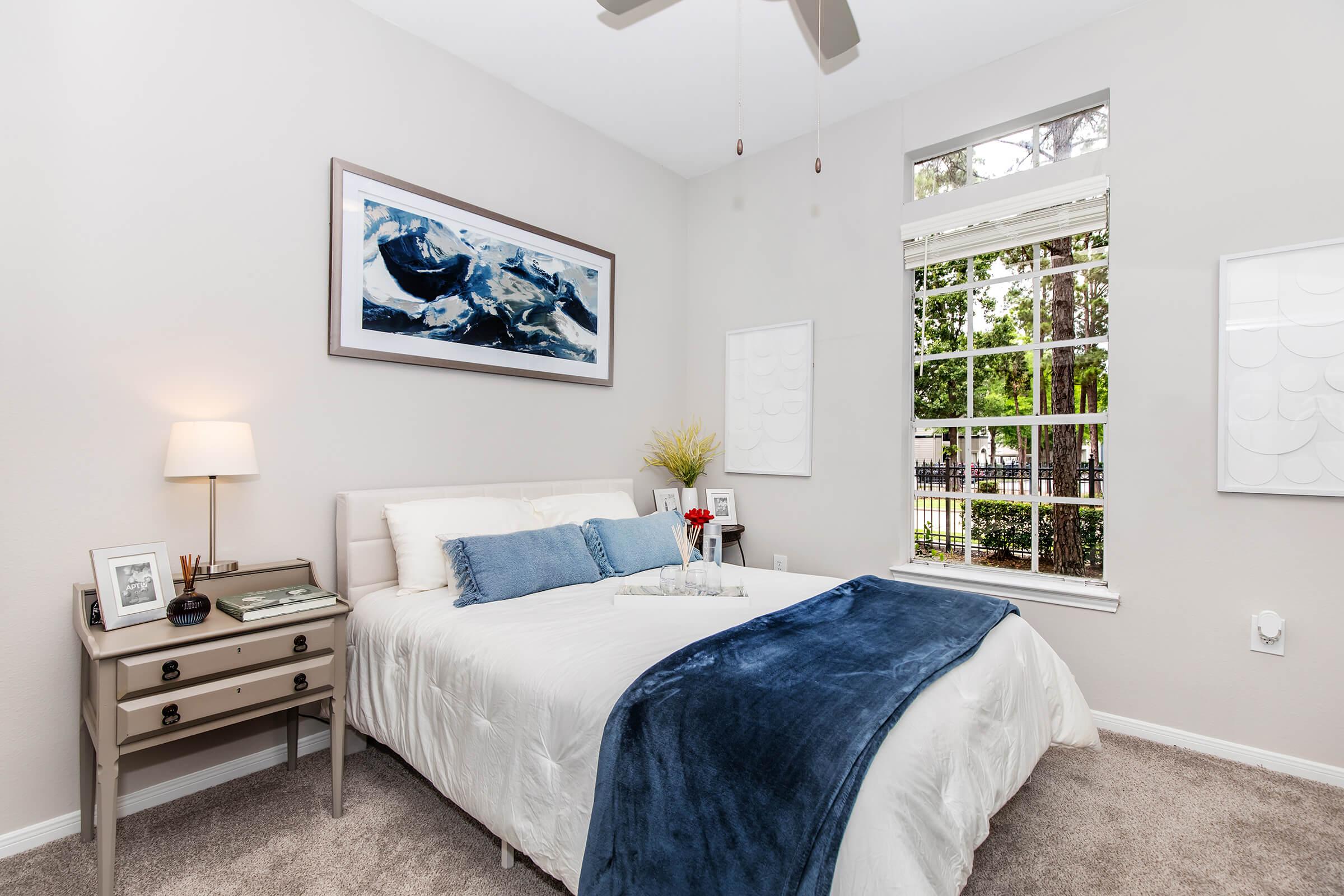 A cozy bedroom featuring a bed with white bedding and a blue throw blanket. A bedside table with a lamp and decor items is beside the bed. The room has light gray walls and large windows with white frames, letting in natural light. Decorative artwork hangs above the bed, complementing the serene atmosphere.