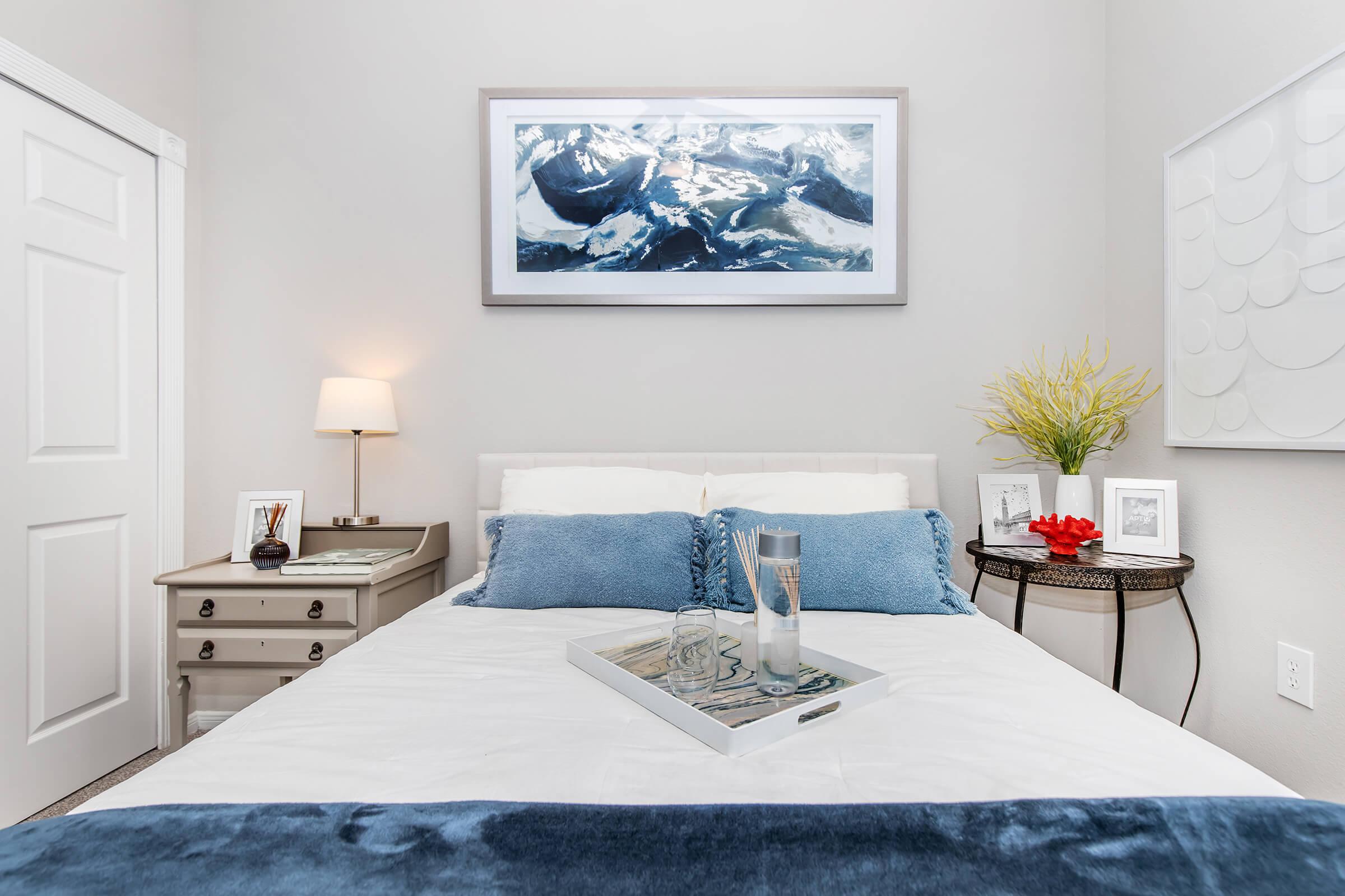 A cozy, well-decorated bedroom featuring a white bed with blue accents, a small bedside table with a lamp, and framed photos. The wall displays a large landscape painting of mountains, and there is a decorative tray on the bed. Greenery adds a touch of color to the room.