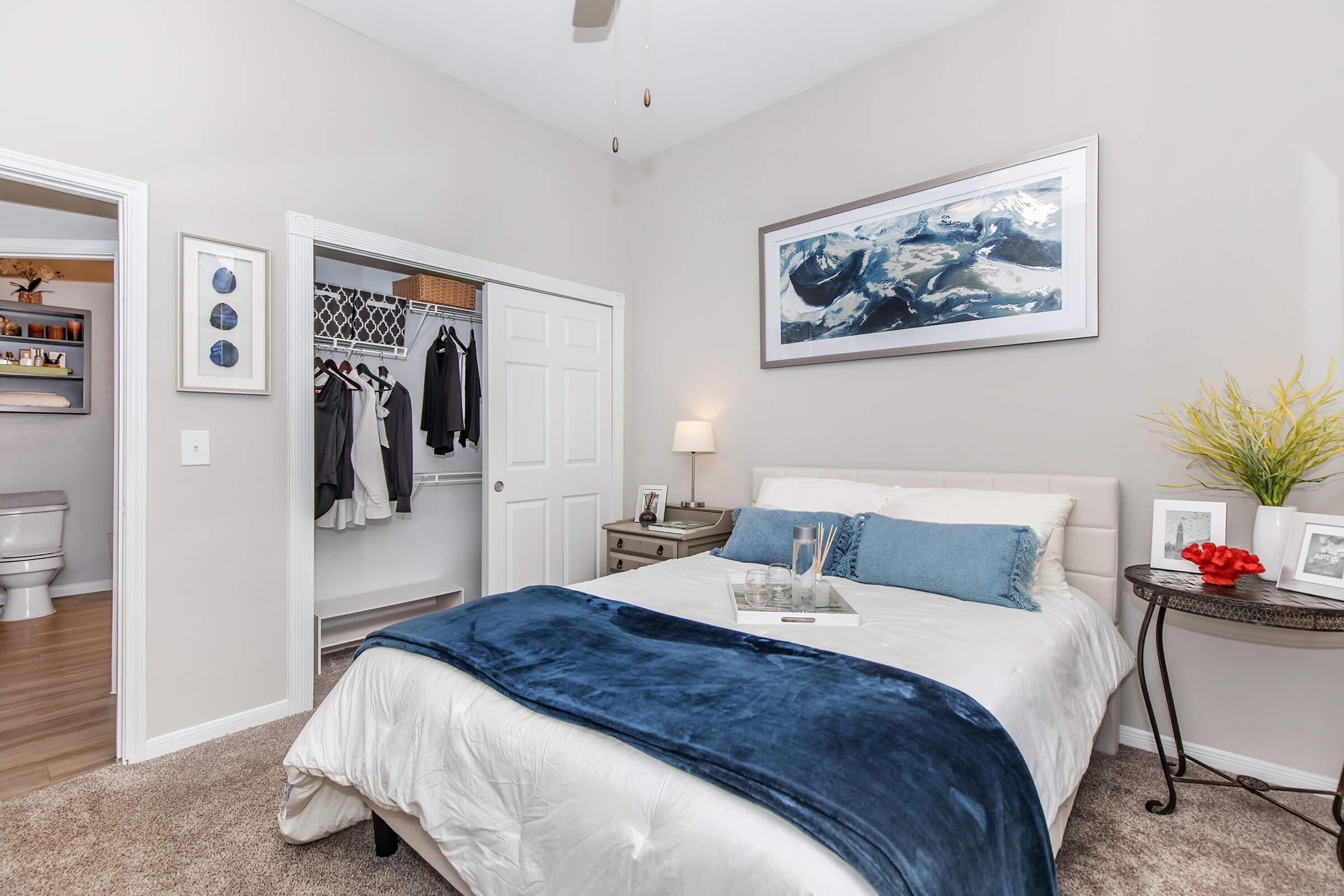 A cozy bedroom featuring a white bed with a blue throw blanket, a nightstand with decorative items, and a painting of mountains on the wall. A spacious closet is visible, and a doorway leads to a bathroom. The room has neutral-colored walls and carpet, creating a warm and inviting atmosphere.