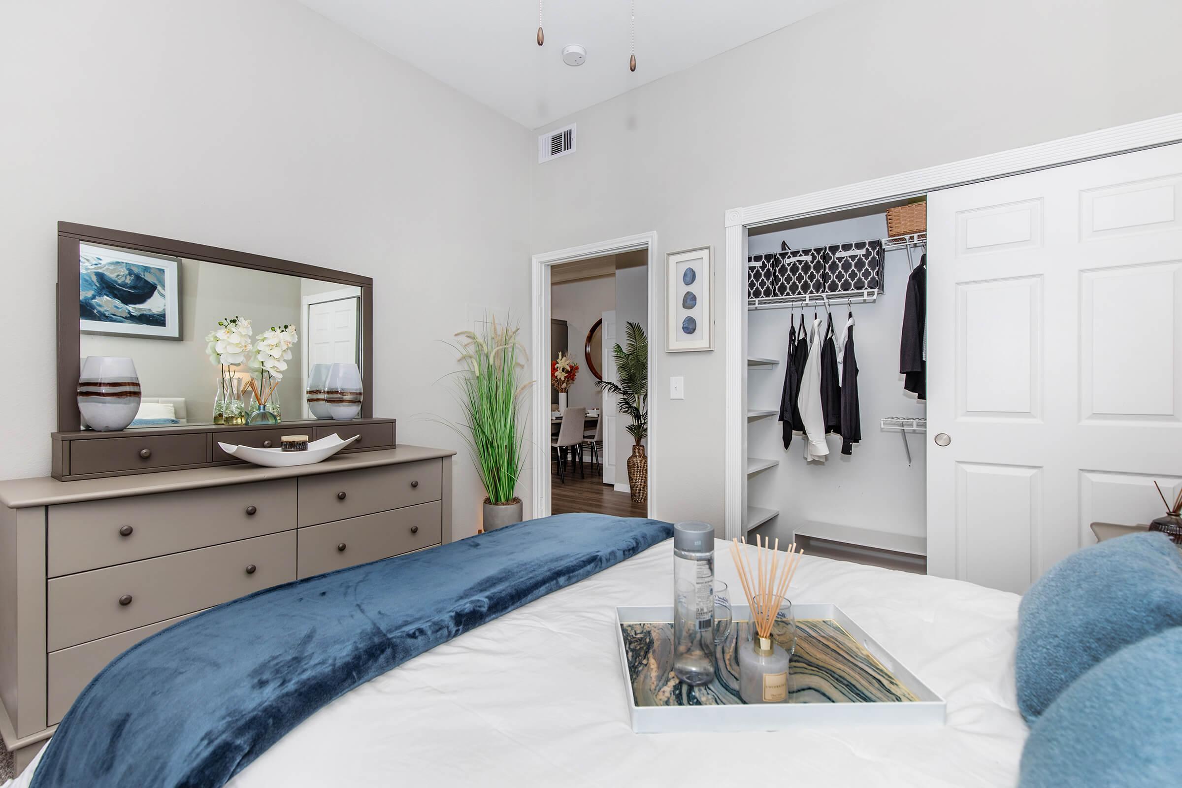 A well-decorated bedroom featuring a gray dresser with a mirror, a neatly made bed with blue bedding, and a decorative tray. In the background, there’s a closet with clothes hanging and plants adding a touch of greenery. The walls are painted in a light color, creating a bright and airy atmosphere.