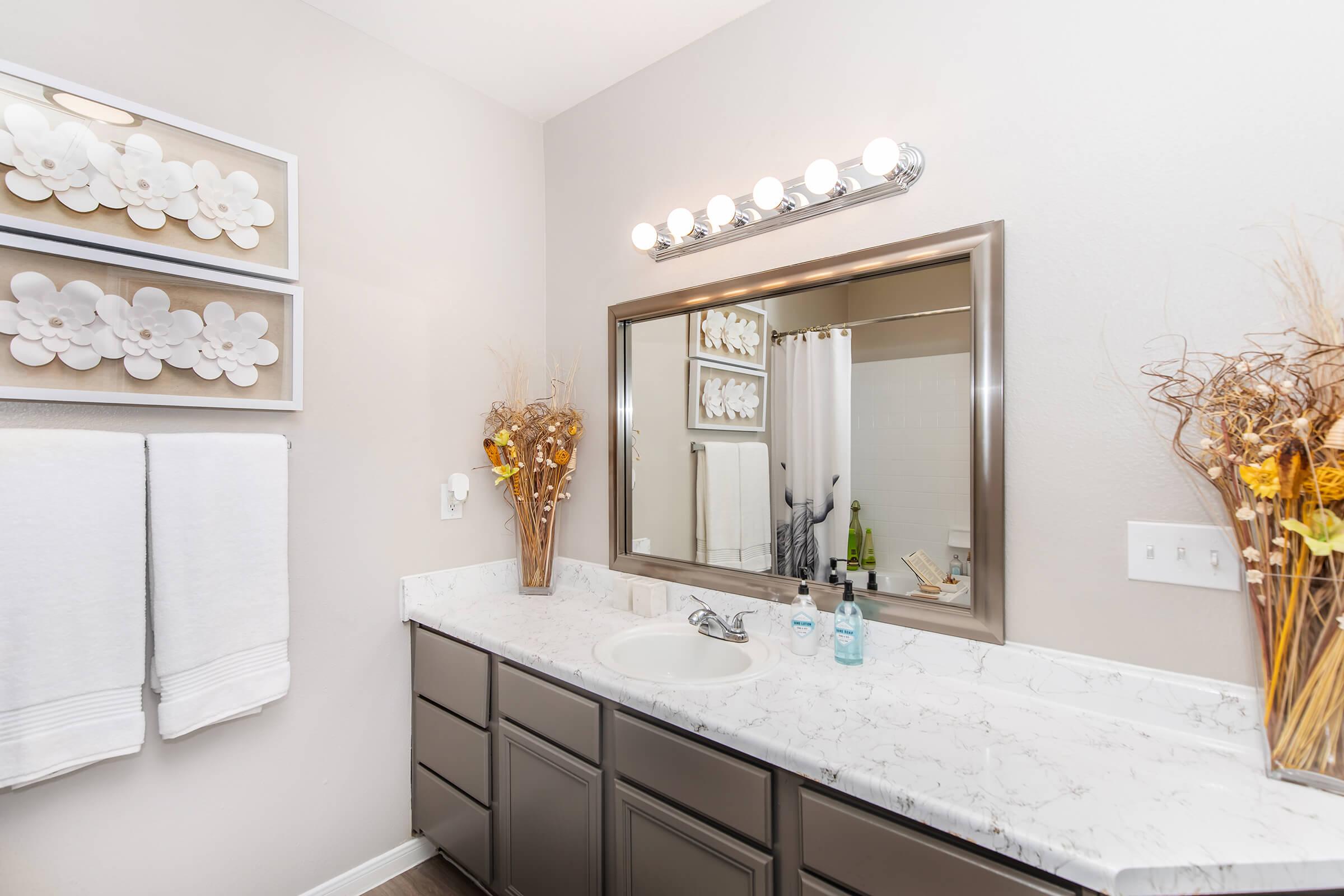 A modern bathroom featuring a double vanity with a marble countertop, a large mirror with lighting above, and two white towels hanging. Decor includes flower arrangements in vases and wall-mounted shelves with decorative items, creating a clean and stylish aesthetic.
