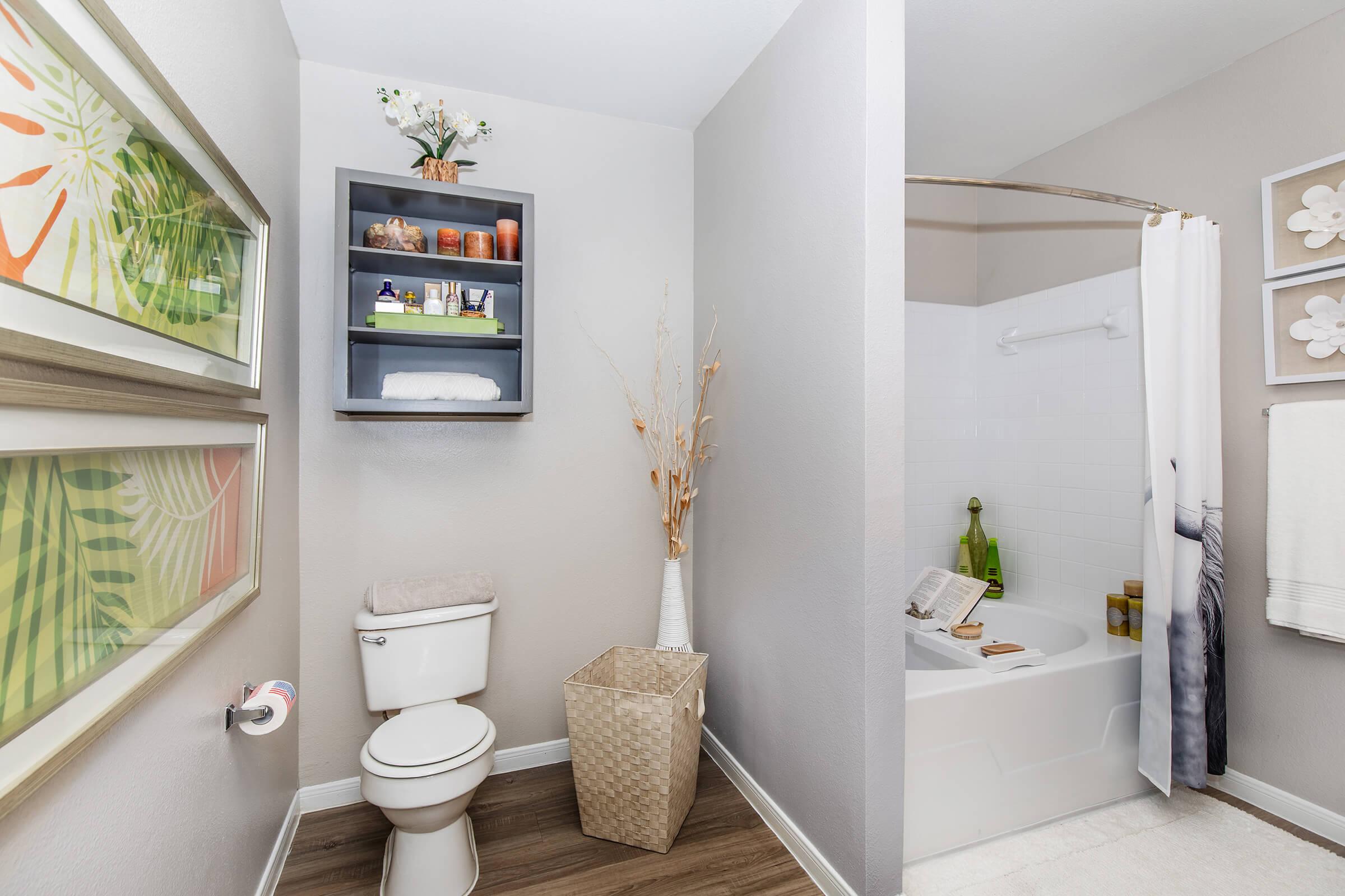 A clean, modern bathroom featuring a toilet, a decorative shelving unit with toiletries, and a bathtub with a shower curtain. Wall art with tropical themes is displayed. A woven basket sits on the floor, and a small vase with dried flowers adds a touch of decor.