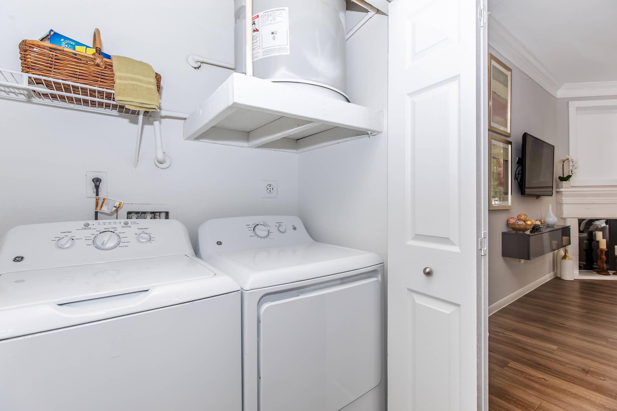 A laundry room featuring a stackable washer and dryer unit. There are shelves above with a basket and cloth items. The room has a door leading to another space that includes a television and decorative items. The flooring is wooden, and the walls are a light color.