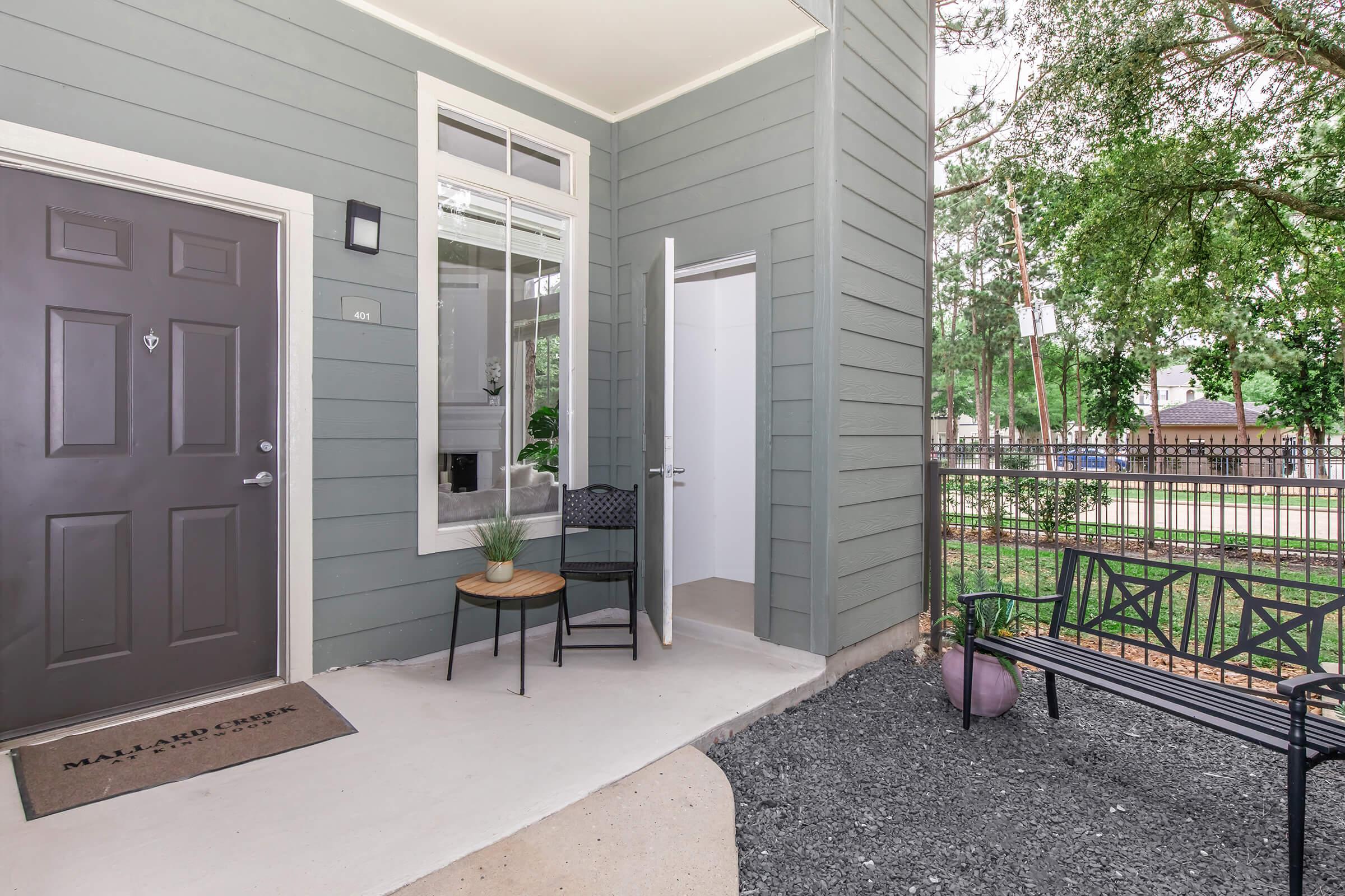 A modern entryway featuring a gray front door with a small welcome mat, a potted plant on a circular table, and a chair beside it. An open interior door leads to a hallway, and outside, there's a black bench and gravel landscaping. Trees line the background, enhancing the inviting atmosphere.