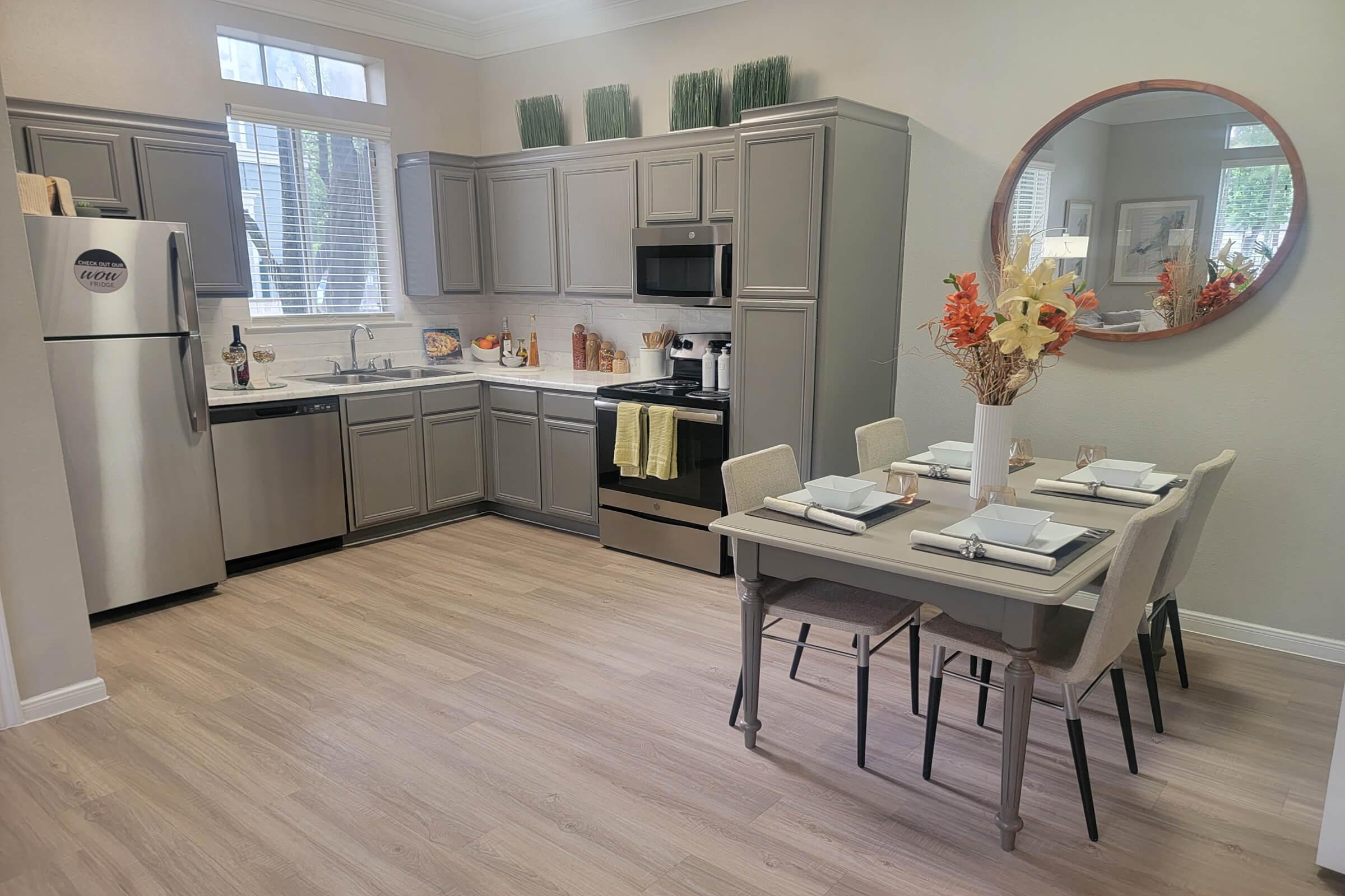 Modern kitchen featuring gray cabinetry, stainless steel appliances, and a round mirror. A dining table set for four, with decorative floral arrangements, is positioned adjacent to the kitchen. Natural light streams through a window, highlighting the light wood flooring and warm décor.