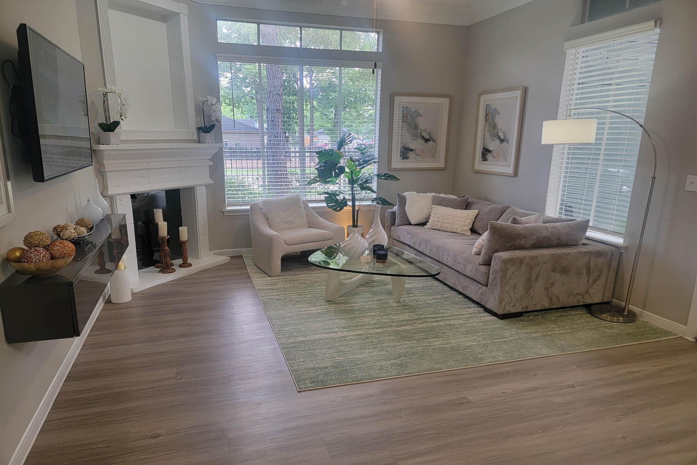 A cozy living room featuring a light gray sofa, a glass coffee table, and a modern floor lamp. There are two chairs beside the sofa and decorative elements like a plant and artwork on the walls. A fireplace is located on the left, and large windows provide natural light, showing a view of trees outside.