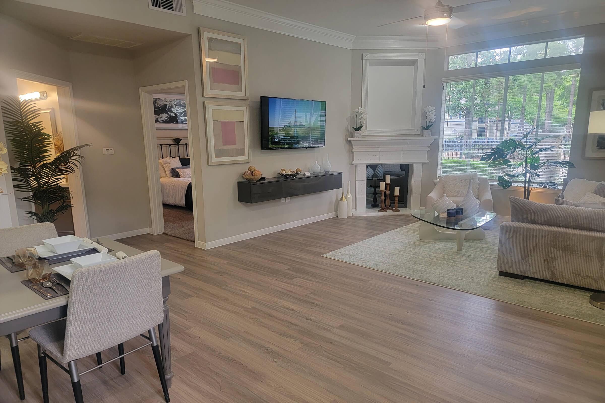 A modern living space featuring a dining area with a table and chairs, a cozy living room with a sofa and decorative pillows, a mounted TV, and a fireplace. Large windows allow natural light, and greenery is visible outside. The decor is stylish and contemporary, with neutral tones and decorative elements throughout.