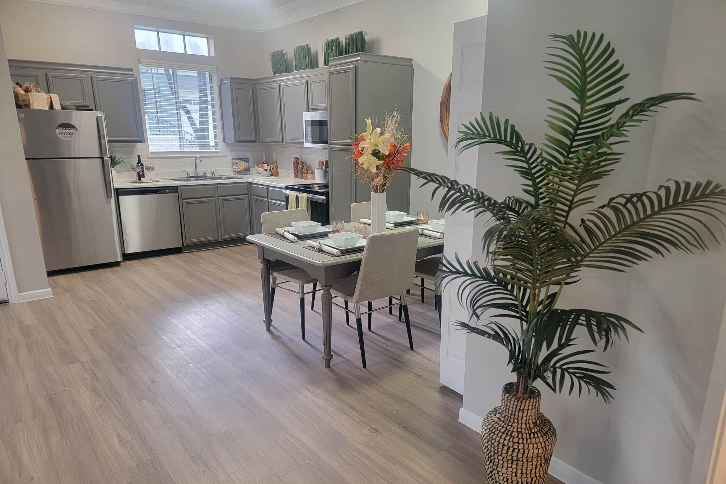 Modern kitchen with gray cabinets and stainless-steel appliances. A dining table set for four, featuring plates and decorative flowers, is positioned centrally. In the foreground, a potted palm adds greenery, while light wood flooring enhances the bright, airy atmosphere. Natural light streams in through a window.