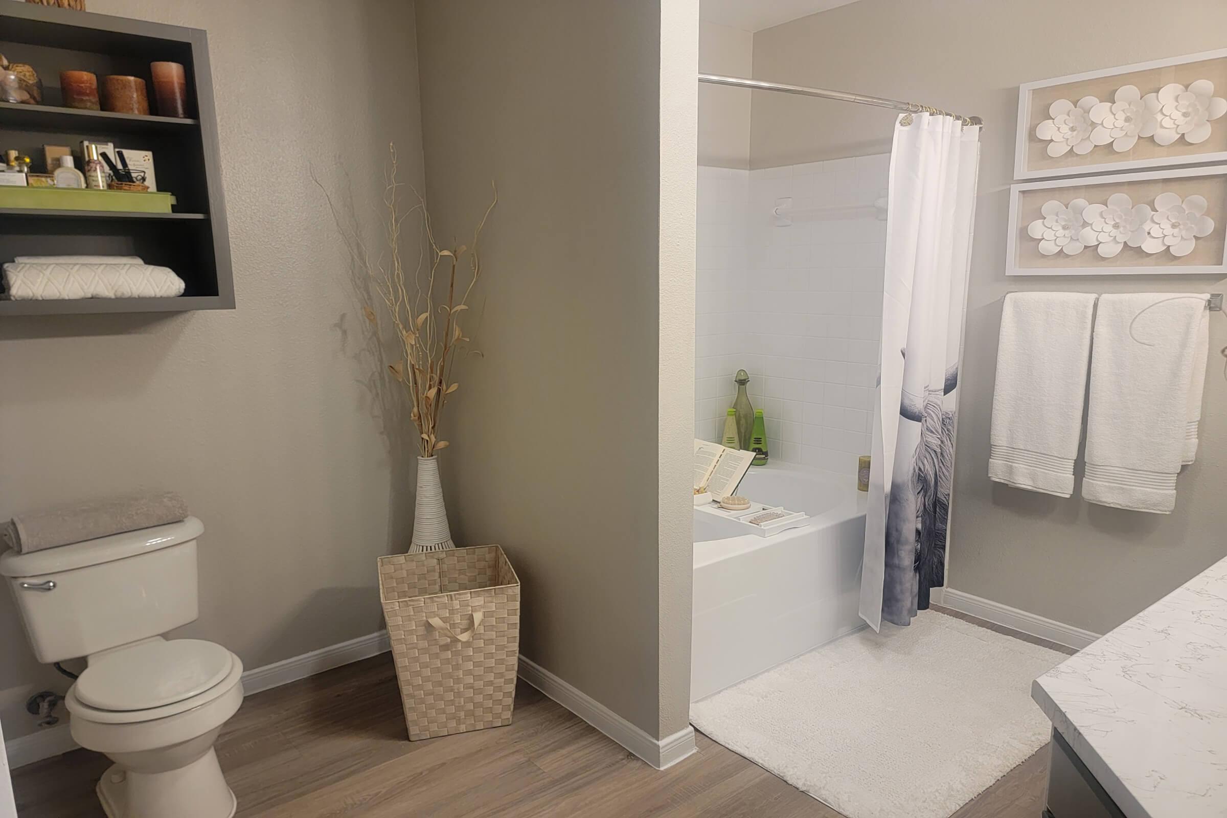 Modern bathroom setup featuring a white bathtub with a shower curtain, a toilet, and a laundry basket. The space includes decorative accents like a shelf with toiletries, a vase with dried plants, and a plush towel display. Light color palette creates a clean and inviting atmosphere.