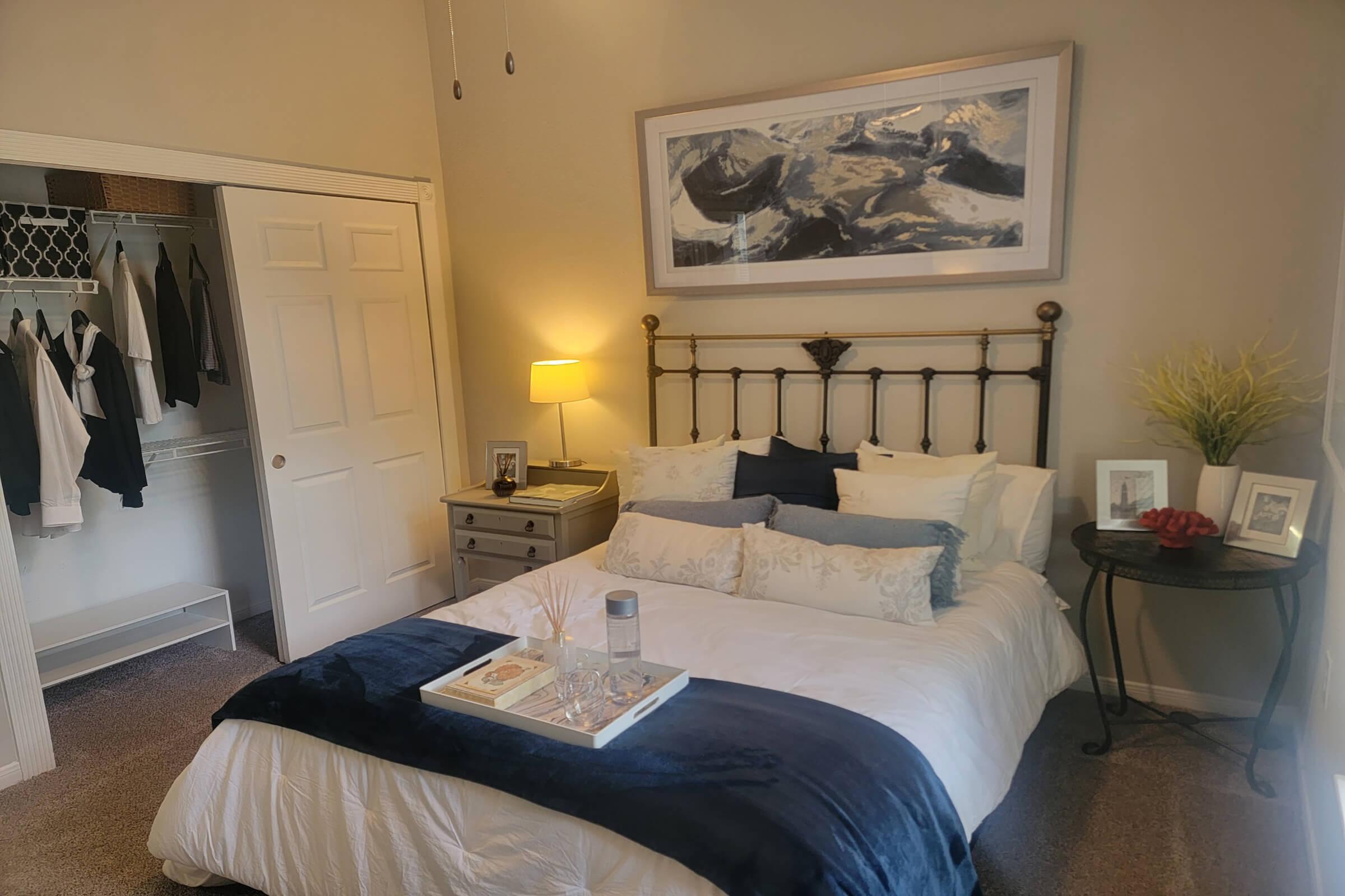 A cozy bedroom featuring a queen-sized bed with white and blue bedding, a bedside table with a lamp, and decorative pillows. A framed abstract painting hangs above the bed. A closet is visible, containing neatly hung clothing. A small round table next to the bed has a tray with a bottle of water and decorative items.