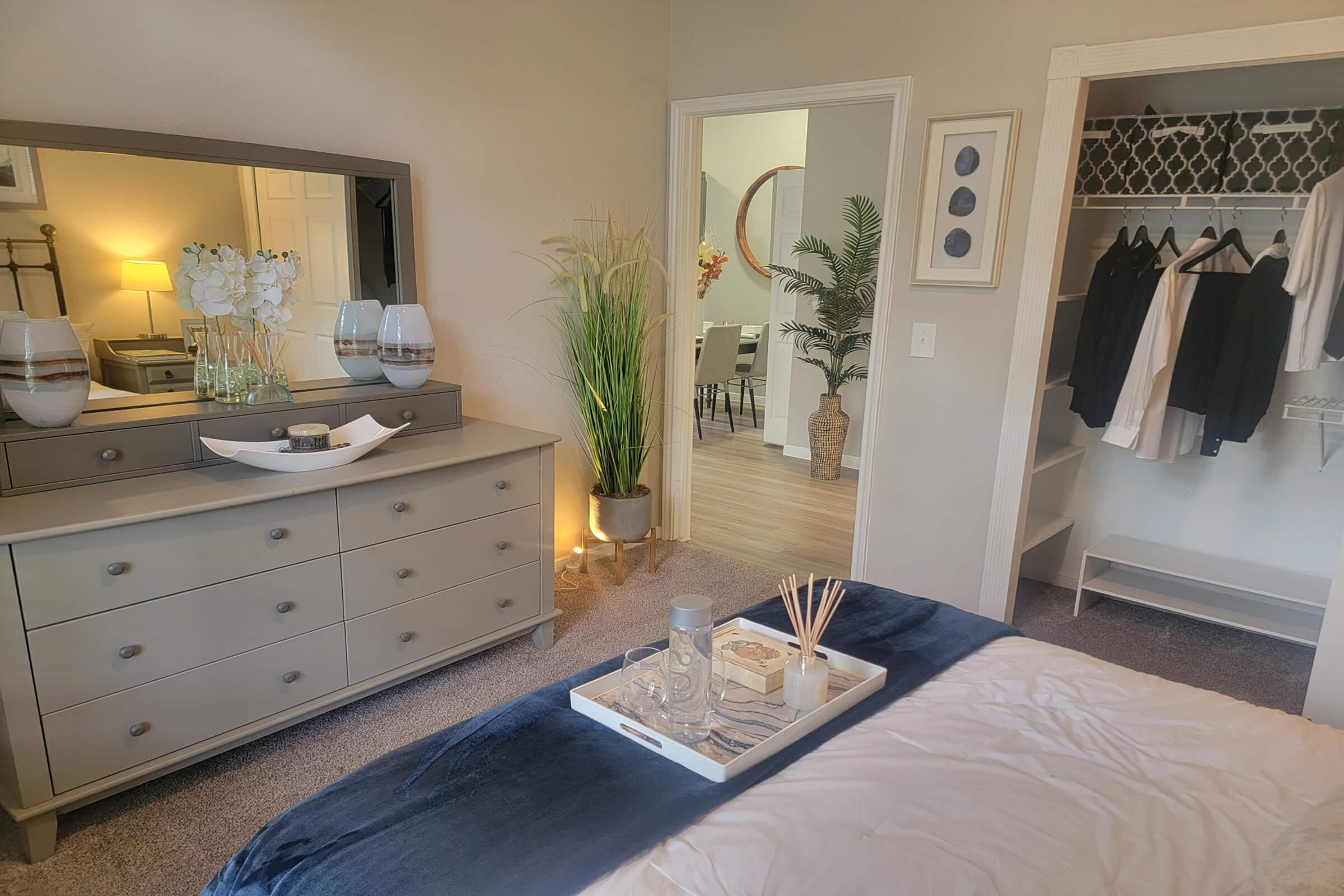 A cozy bedroom featuring a gray dresser with decorative items, a large mirror, and a doorway leading to another room. There are plants, a neatly made bed with a blue throw blanket, and a tray with candles and reed diffusers on the bed. A closet with hanging clothes is visible in the background.
