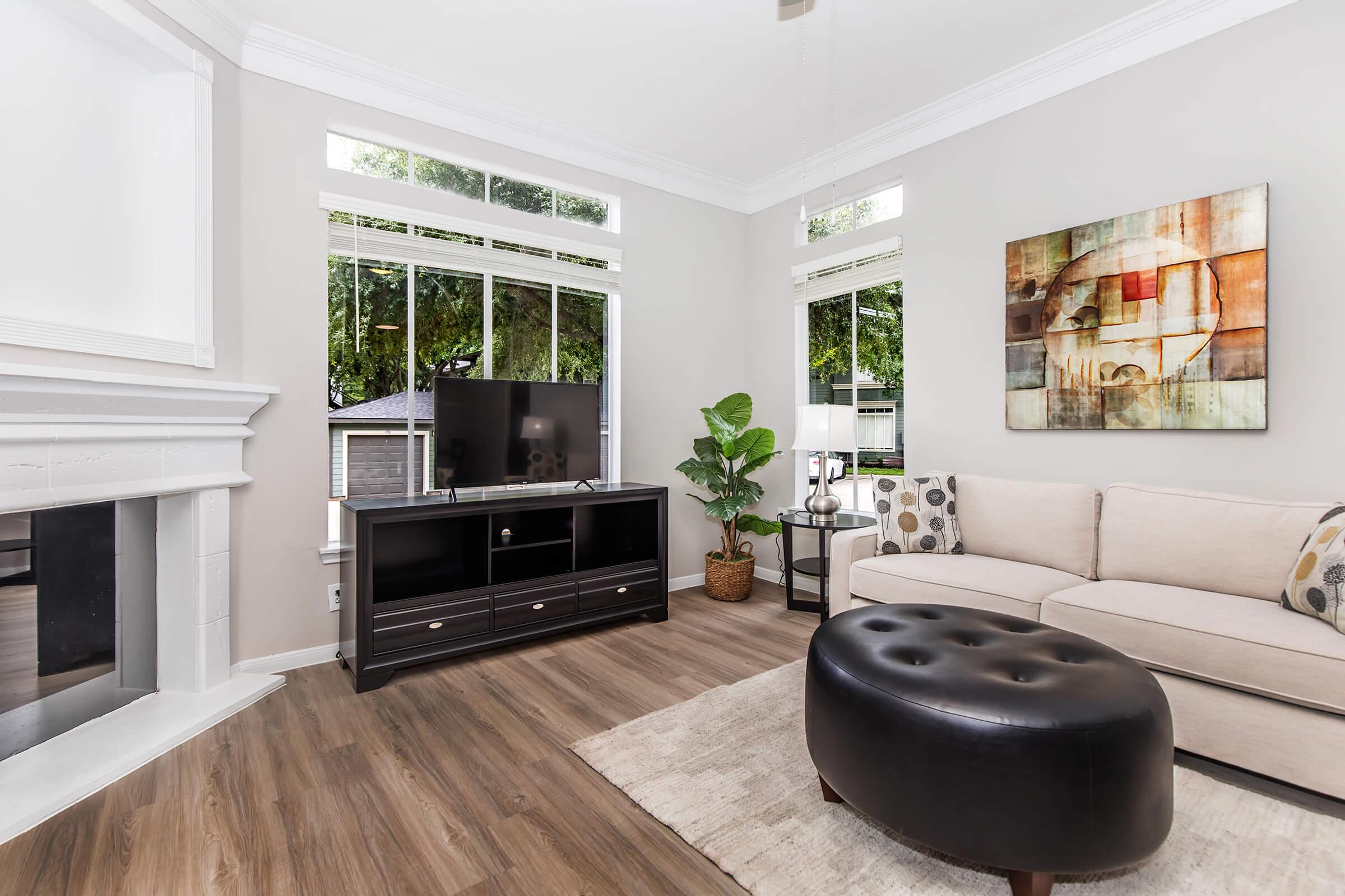 A bright living room featuring a beige sofa with decorative pillows, a round black ottoman, a modern TV stand with a television, and large windows allowing natural light. A potted plant adds a touch of greenery, and a wall art piece is displayed above the sofa. The floor is made of wooden planks.