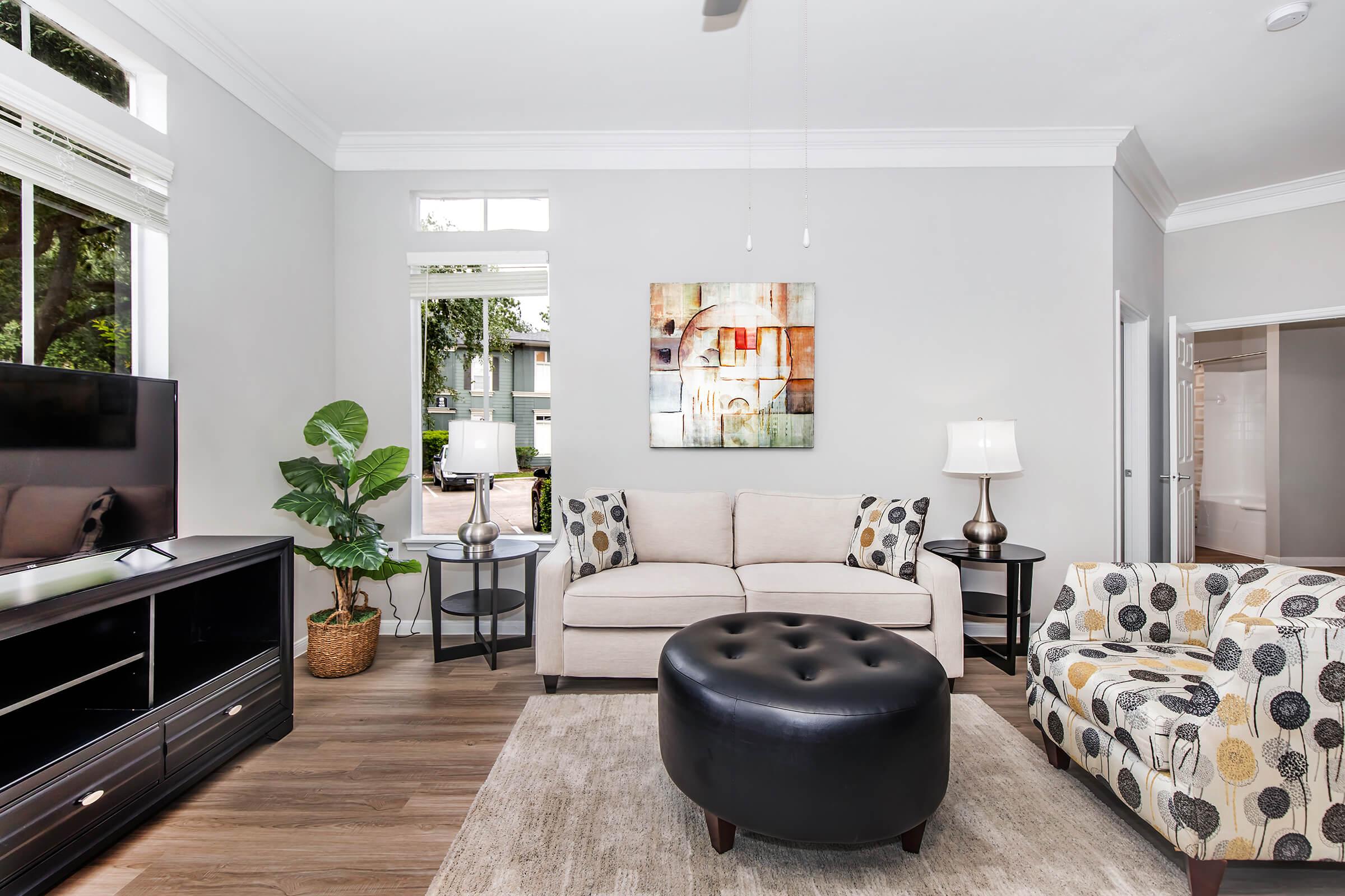 A modern living room featuring a beige sofa, a black tufted ottoman, and a patterned armchair. There is a television on a dark wood stand, a decorative plant in a woven basket, and two lamps on side tables. A colorful abstract painting hangs on the wall, adding a touch of art to the space. Large windows allow natural light to fill the room.