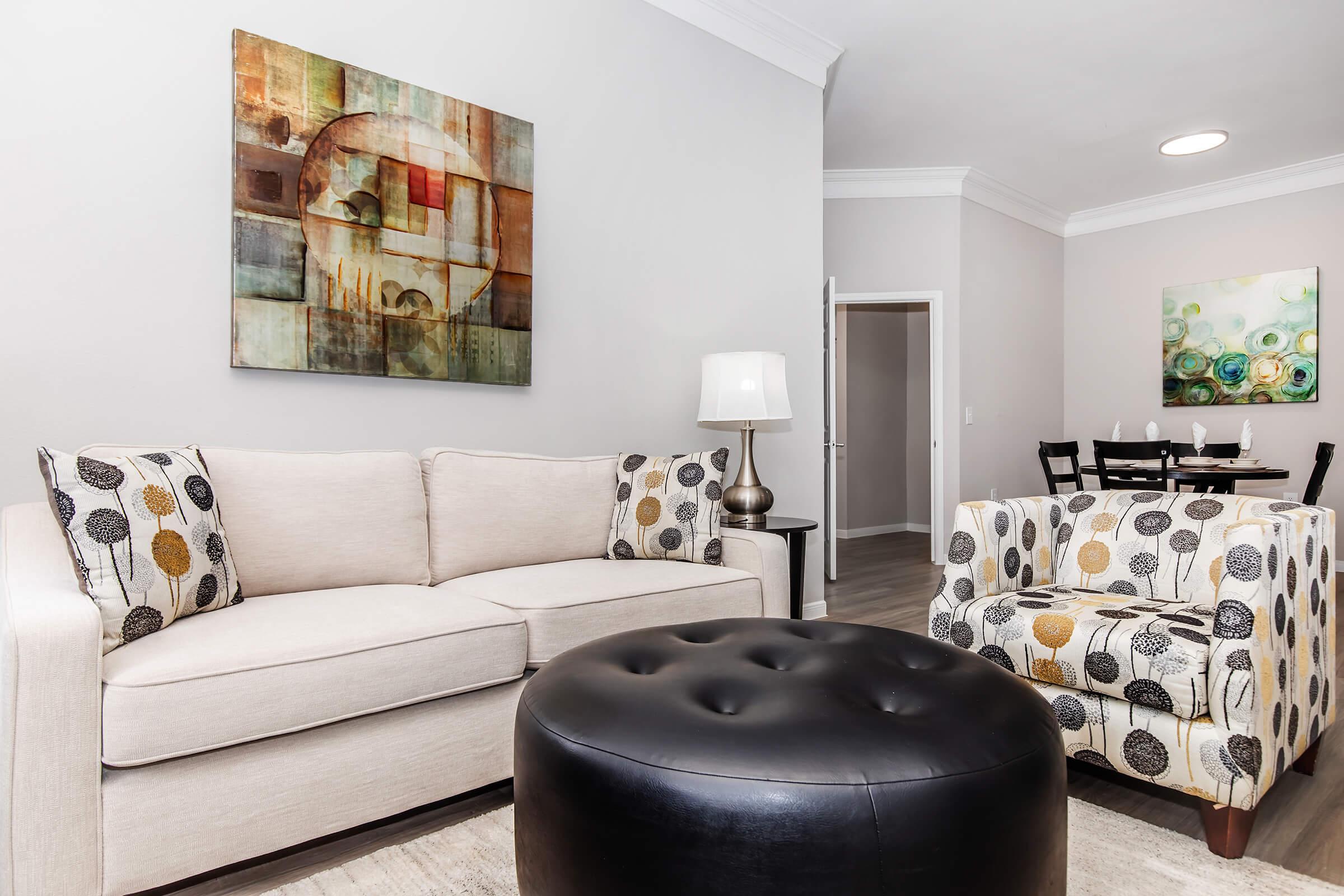A modern living room with a light beige sofa adorned with patterned throw pillows, a round black ottoman, and an accent chair featuring a floral design. Artwork hangs on the pale gray walls, complementing the space. A dining area with a round table and chairs is visible in the background.