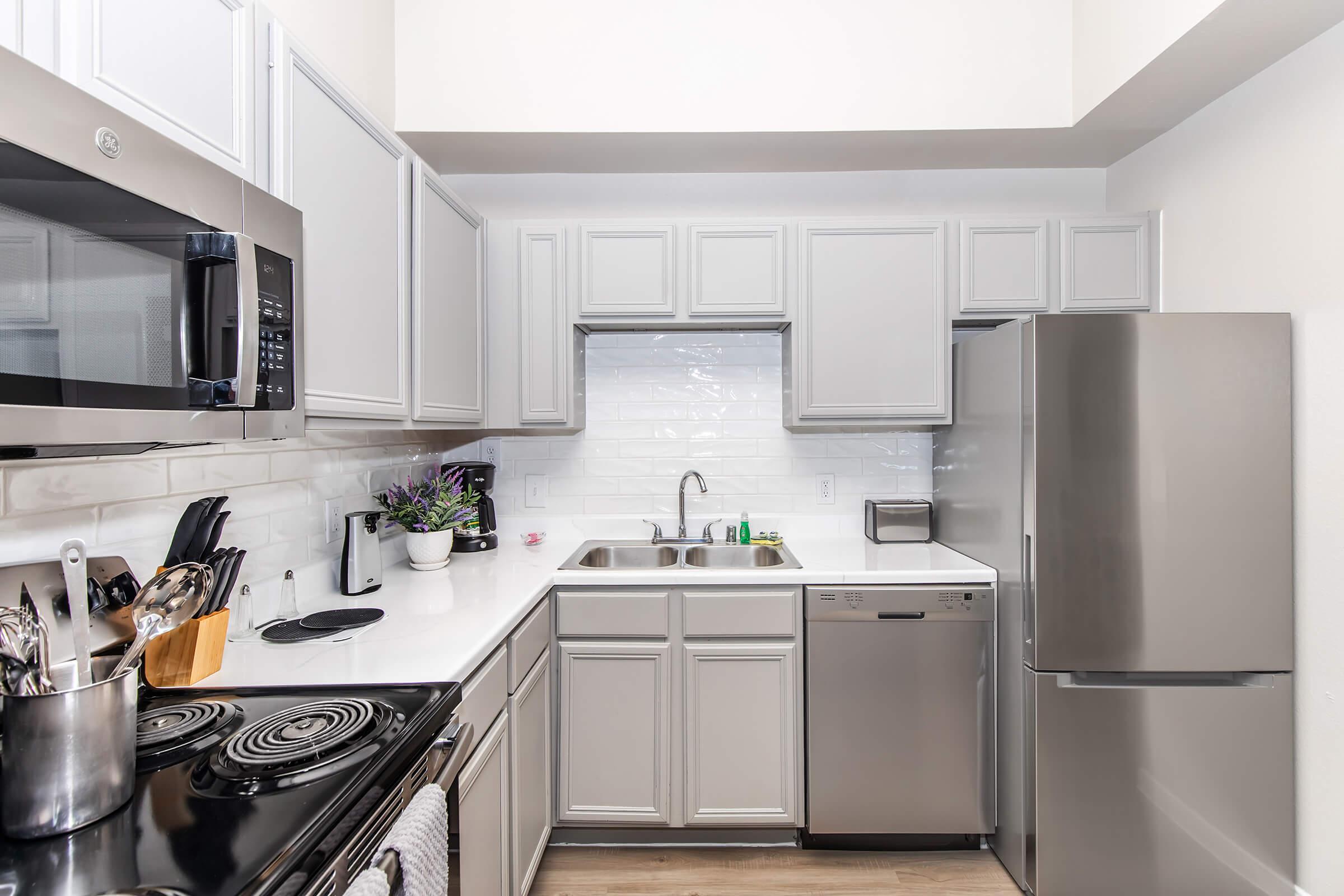 A modern kitchen featuring gray cabinetry, stainless steel appliances including a microwave, dishwasher, and refrigerator. A white countertop and backsplash enhance the bright space, while various kitchen utensils and a small potted plant add decorative touches.
