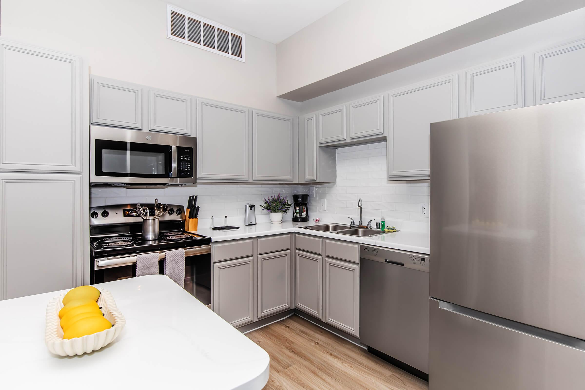Modern kitchen featuring gray cabinets, stainless steel appliances including a microwave, stove, dishwasher, and refrigerator. A white countertop with a decorative dish of yellow lemons adds a pop of color. The space has a clean, organized look with a minimalist design.