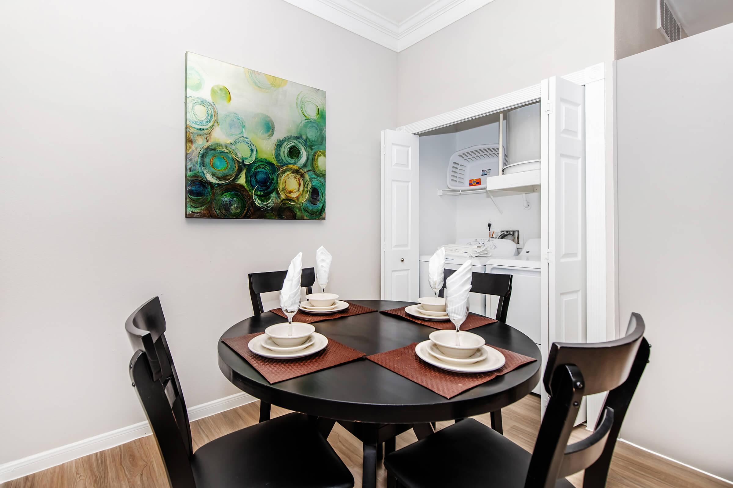 A dining area featuring a round black table set with plates and napkins, surrounded by four chairs. A colorful abstract painting is mounted on the wall, and a laundry area is visible through an open door, showing a stacked washer and dryer. The walls are light-colored, creating a bright atmosphere.
