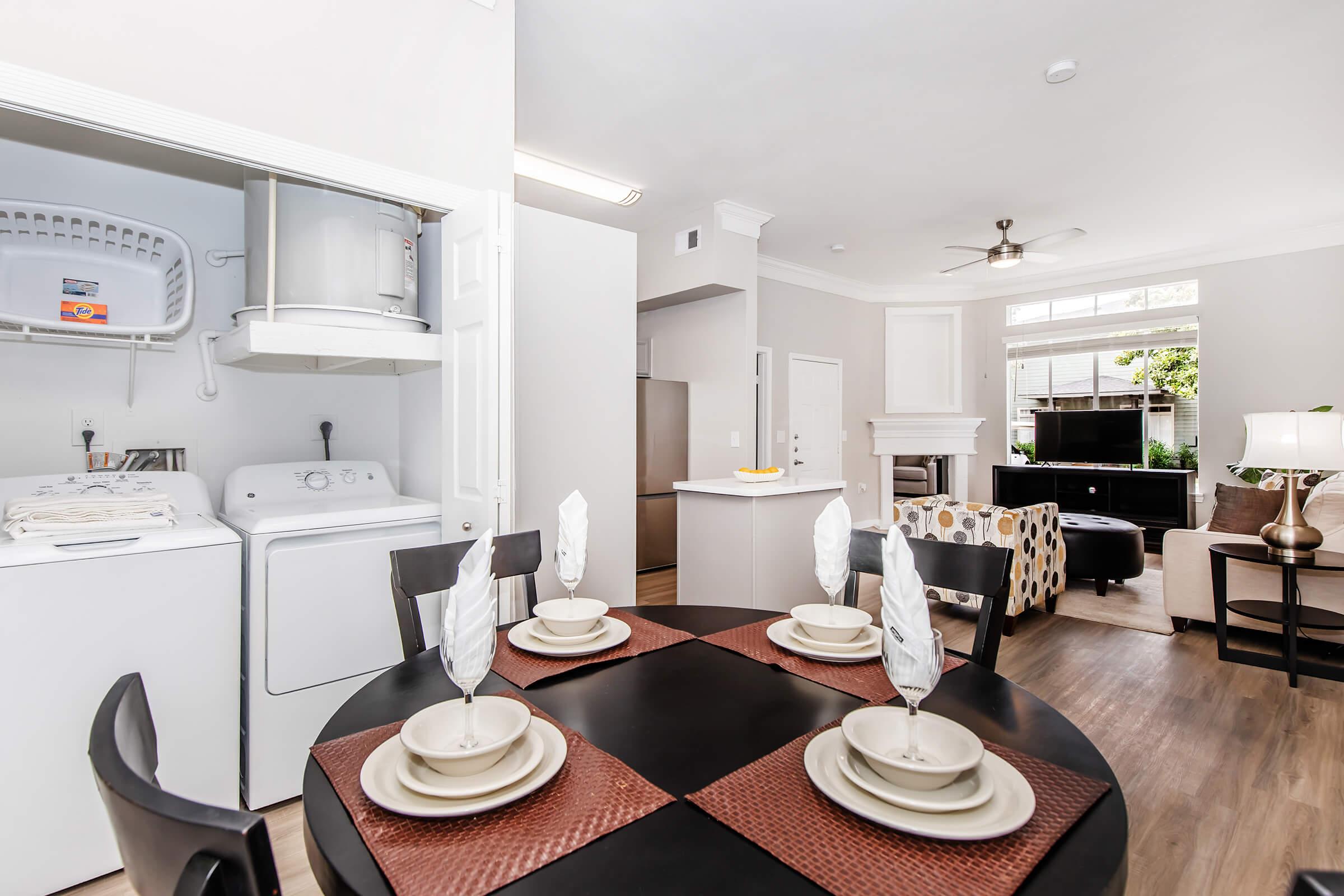 Modern living space featuring a dining area with a round black table set for four, including plates and napkins. In the background, there's a laundry area with a washer and dryer, a well-lit kitchen, and a cozy living room with a black sofa and decorative armchairs. Natural light streams in through large windows.