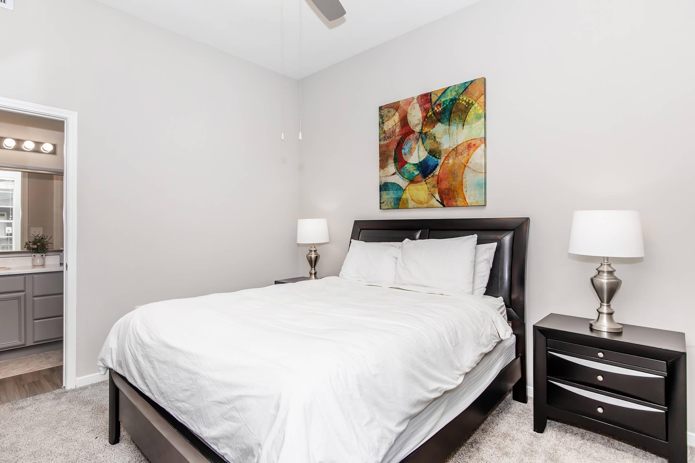 A cozy bedroom featuring a queen-sized bed with white bedding, two bedside tables with silver lamps, and an abstract wall art piece above the headboard. The walls are painted in a light color, and there's a doorway leading to another room, adding a bright and airy feel to the space.