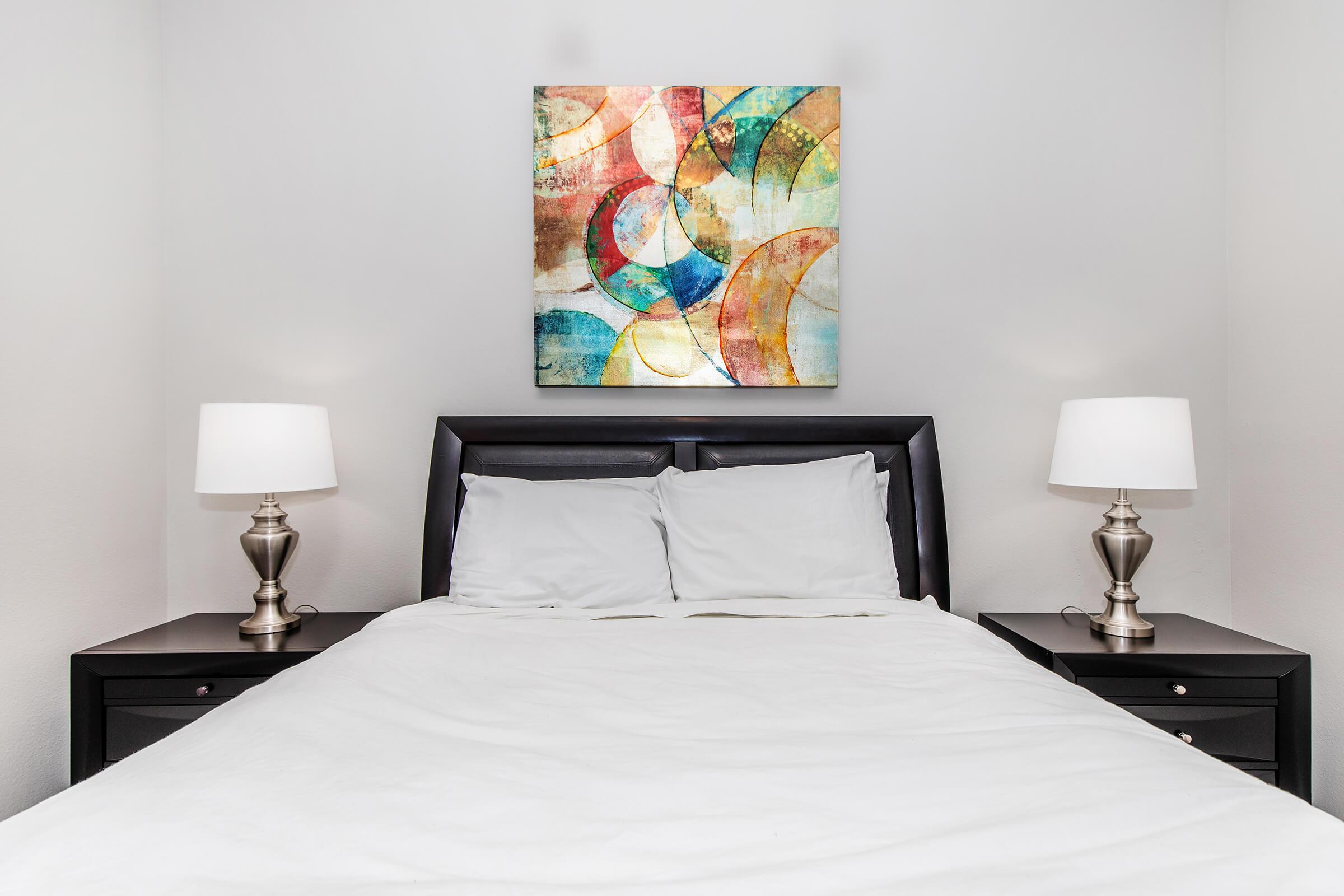 A cozy bedroom featuring a neatly made bed with white linens, flanked by two modern lamps on black nightstands. Above the bed, there is an abstract painting with colorful circular designs, adding a vibrant touch to the serene space. The walls are painted in a light color, enhancing the bright atmosphere.