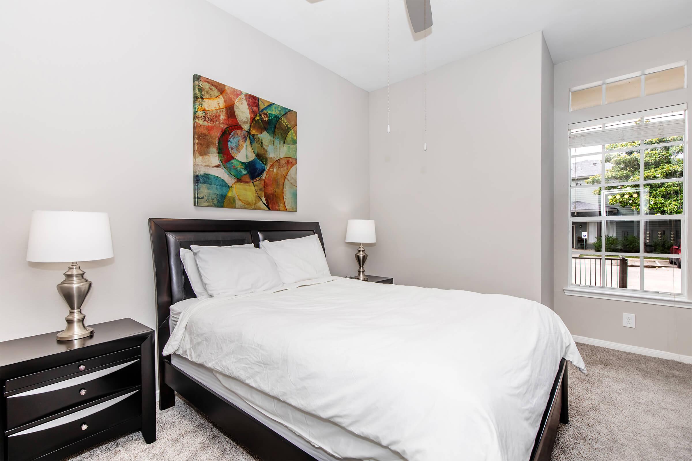 A well-lit bedroom featuring a large bed with white bedding, two bedside tables with lamps on either side, and a colorful abstract painting on the wall. A window offers natural light and a view of the outdoors. The room has a neutral color scheme with soft carpeting.