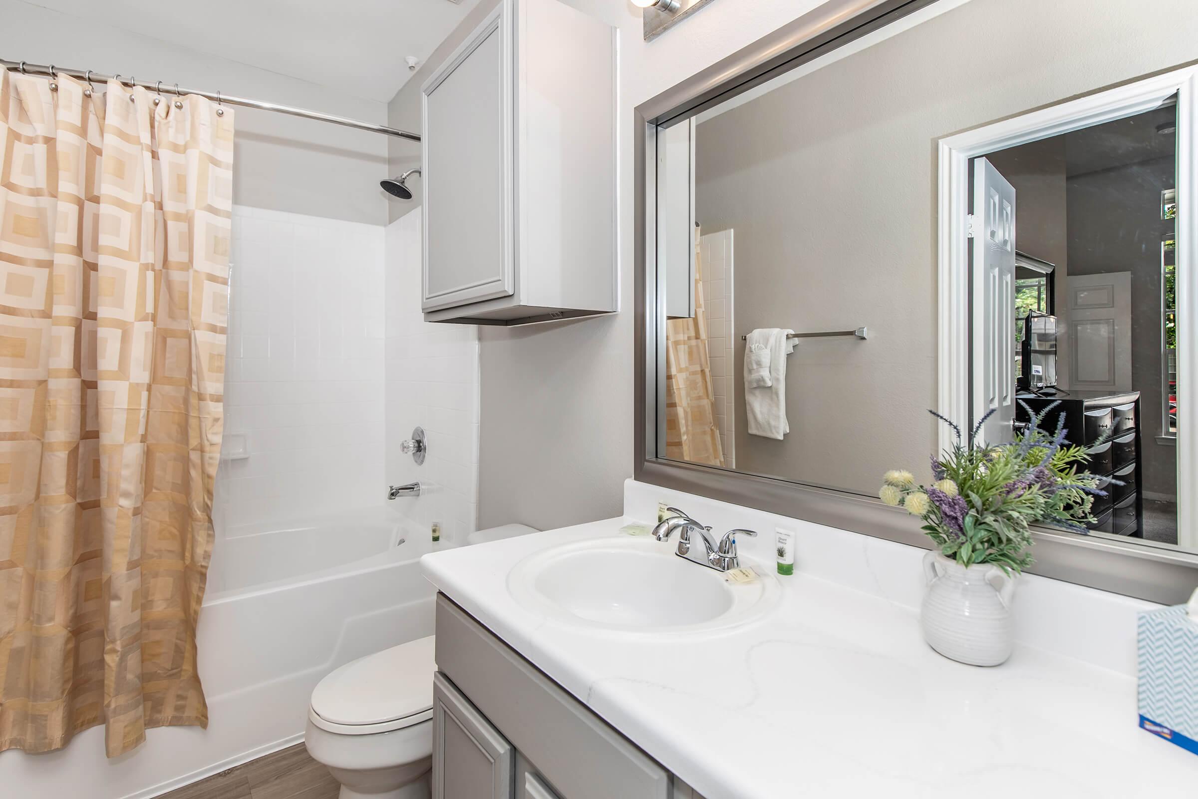 A modern bathroom featuring a white bathtub and shower combination, a light gray cabinet with a sink, a large mirror above the sink, and a cream shower curtain with geometric patterns. A small vase with flowers sits on the countertop, and a towel hangs on a rack. Light-colored walls enhance the bright feel of the space.