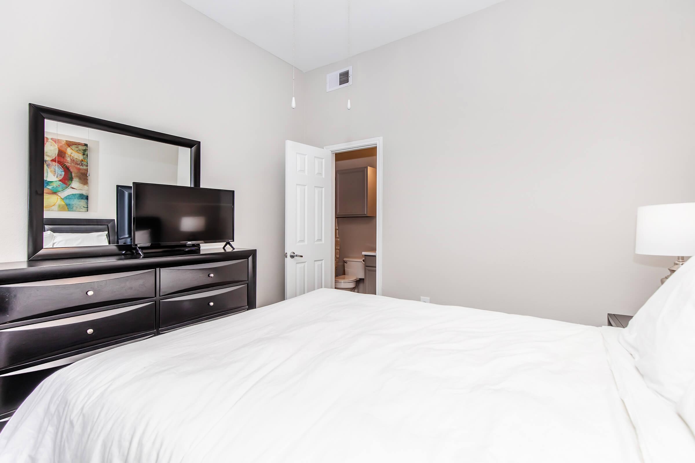 A cozy bedroom featuring a large bed with white linens, a dark wood dresser with a television on top, and a mirror reflecting colorful artwork. A doorway leads to a bathroom in the background, adding a sense of depth to the space. The walls are painted in soft, neutral tones, creating a calm atmosphere.