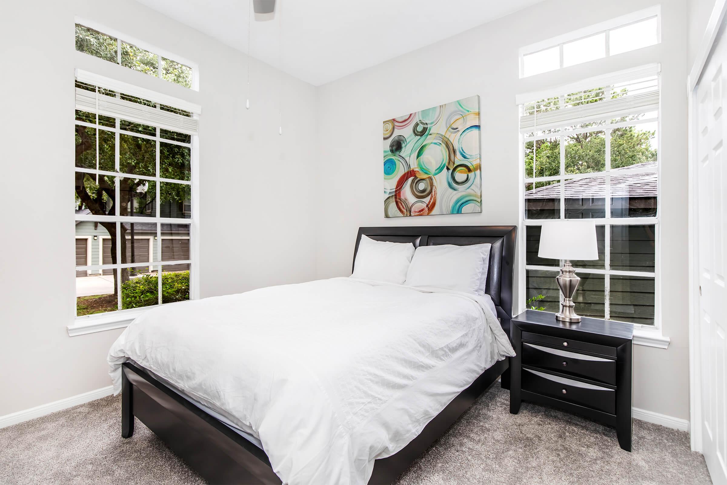 A well-lit bedroom featuring a black bed with white bedding, a nightstand with a lamp, and large windows allowing natural light. The walls are painted in a light color, and a colorful abstract painting is displayed above the bed. The floor is covered with soft carpet, enhancing the cozy atmosphere.
