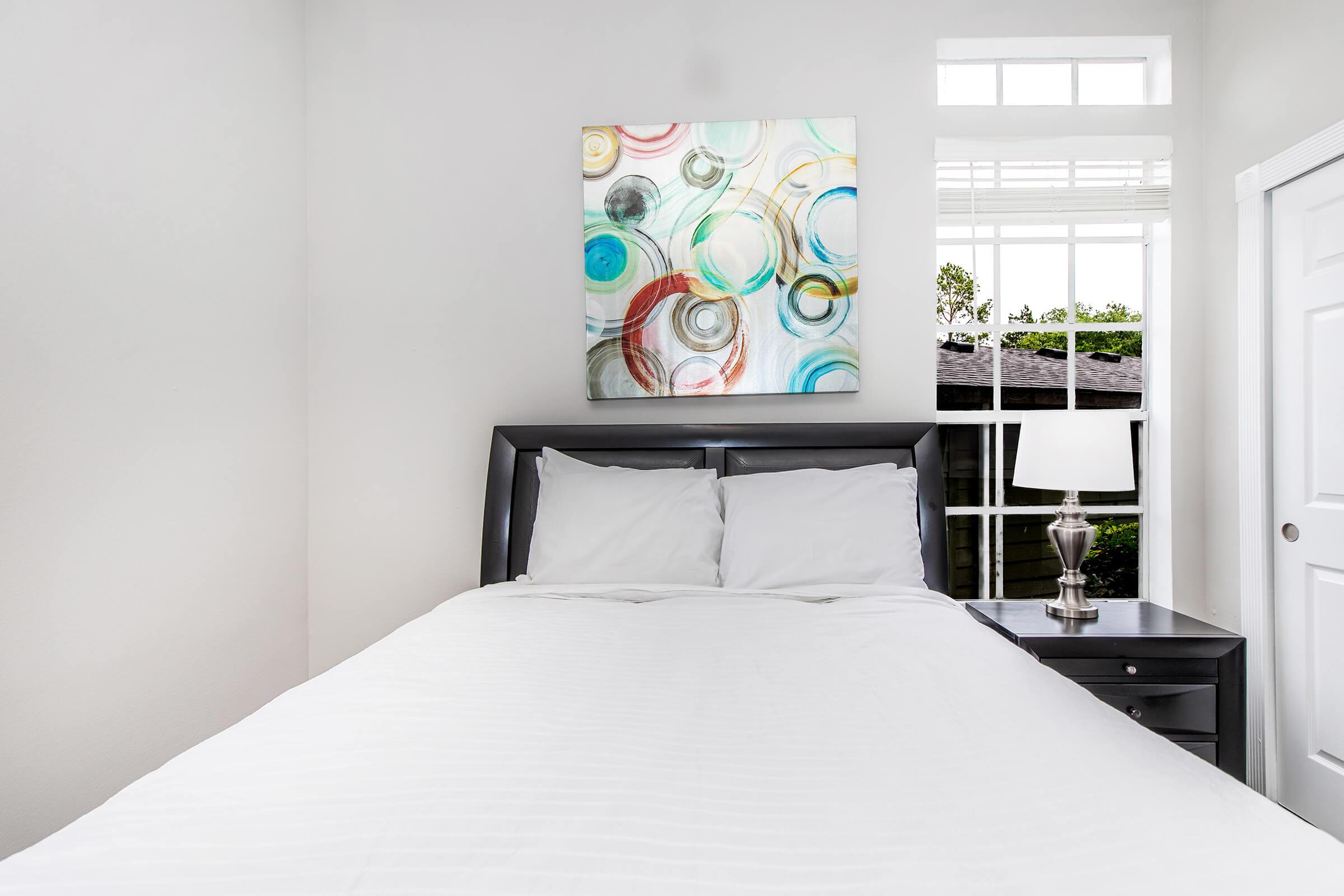 A cozy bedroom featuring a neatly made white bed with two pillows. Above the bed, there's an abstract painting with circular designs in blue, green, and red. A bedside table holds a silver lamp, and a window allows natural light to enter the room, providing a view of greenery outside.
