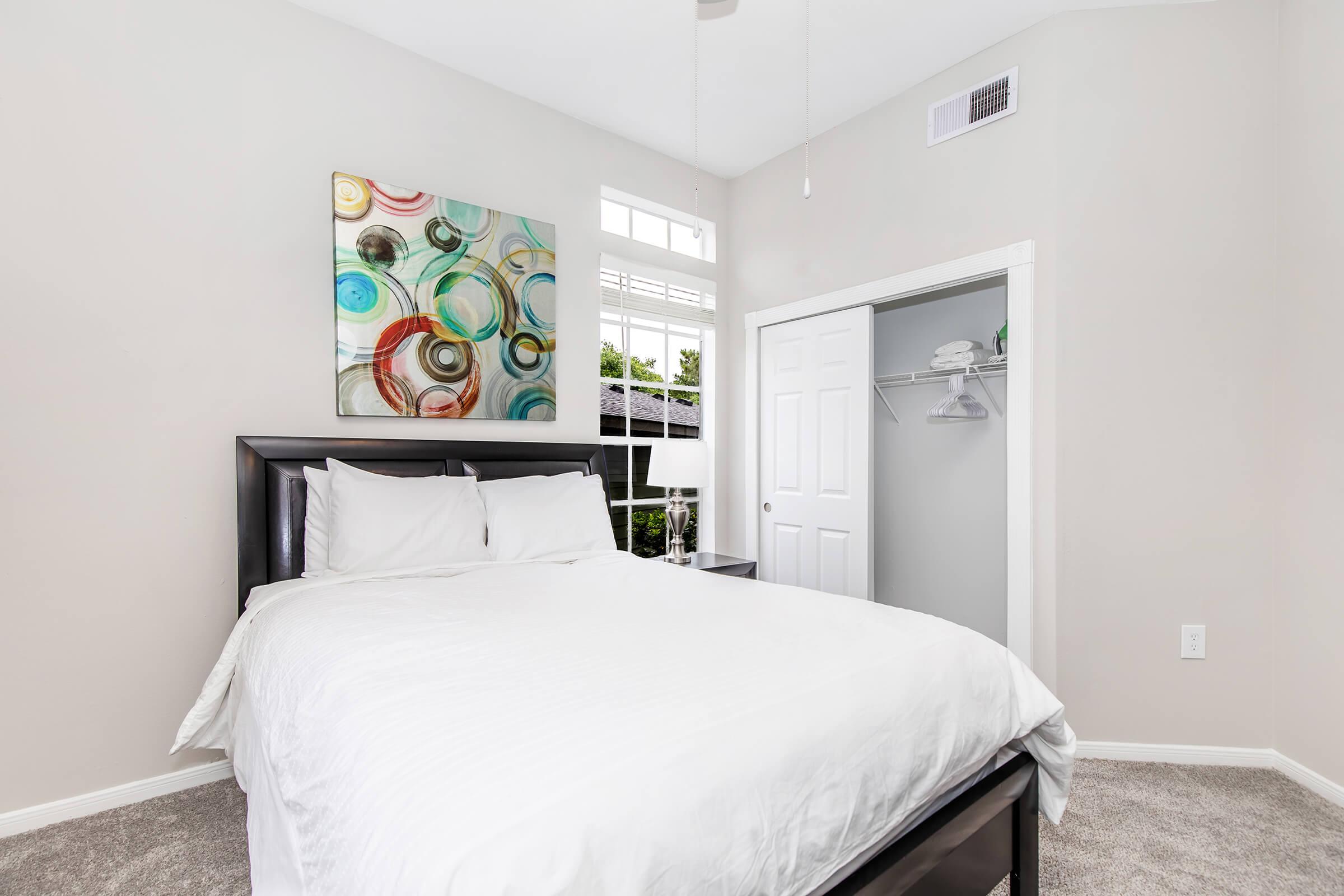 A bright bedroom featuring a cozy bed with white linens, a colorful abstract painting above the headboard, and a closet with a door partially open. The room has neutral walls and a ceiling fan, with natural light streaming in through the windows. Soft carpet adds warmth to the space.