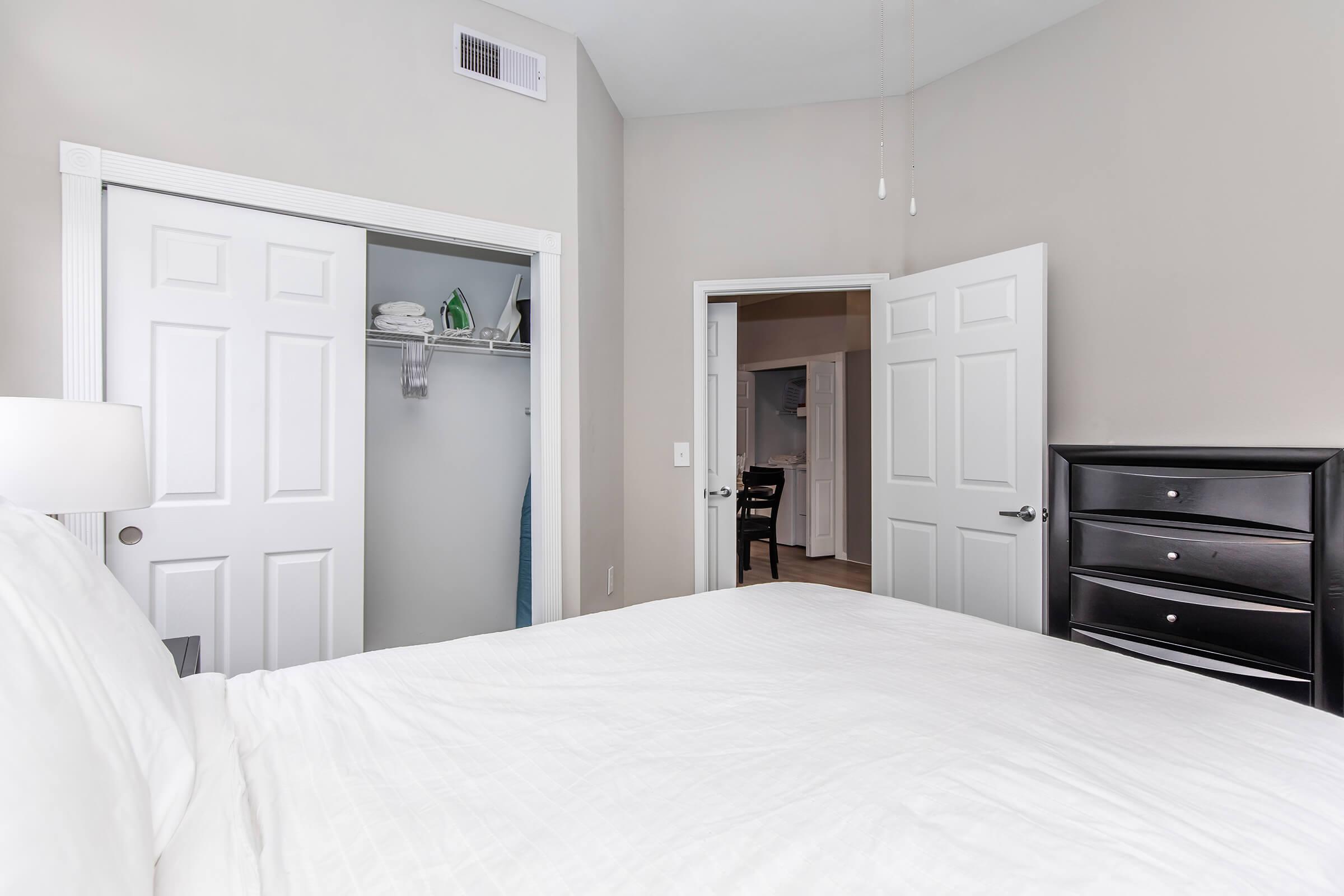 A well-lit bedroom featuring a white bed with a textured comforter. To the left, there is a closed white closet door with a sliver of light visible. In the background, another door leads to a darker room. The room has neutral-colored walls and a black dresser beside the bed.