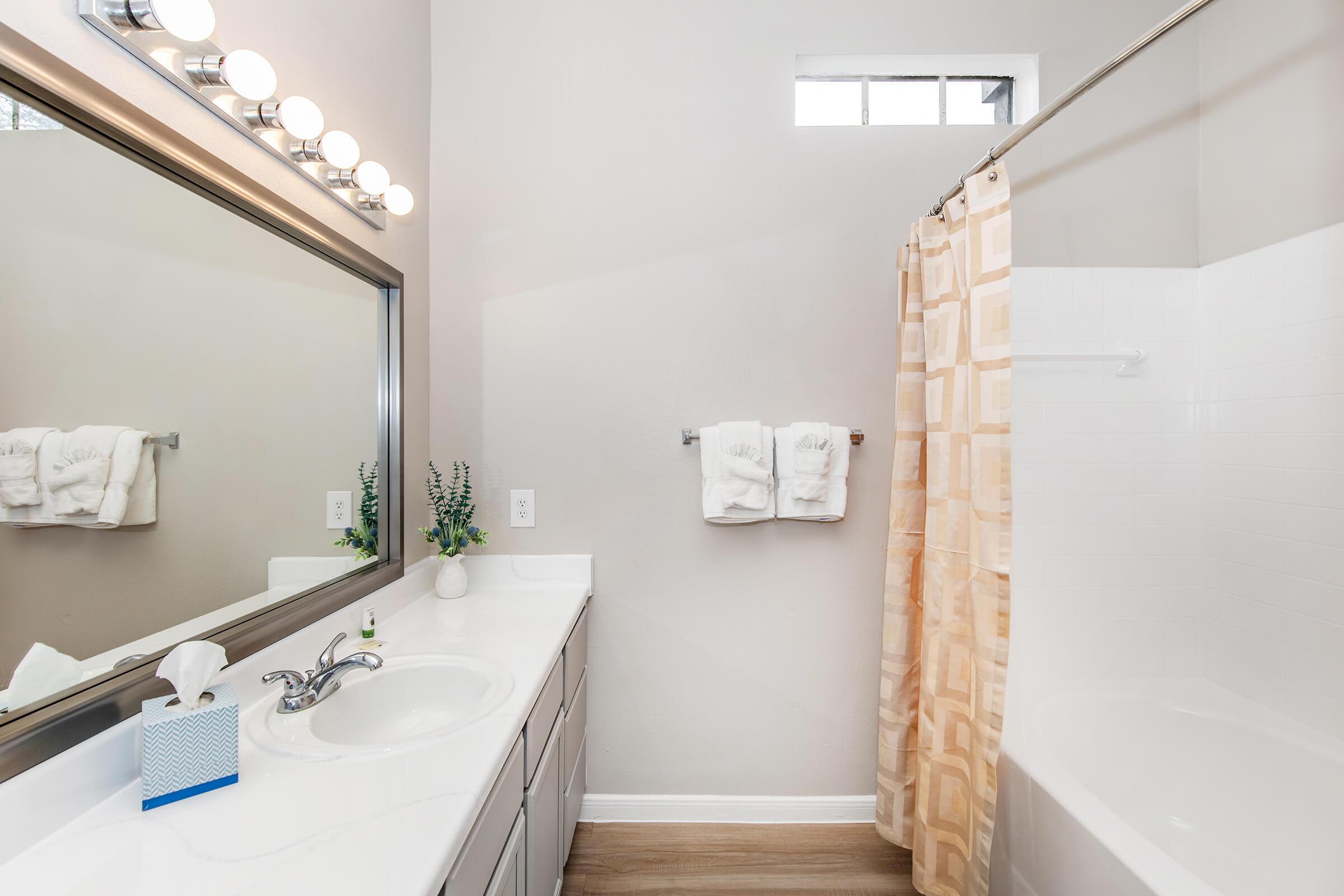 A modern bathroom featuring a white vanity with a mirror above, a silver faucet, and a blue tissue box. On the countertop, there are neatly arranged towels and a small plant. A shower area with a beige curtain and a bathtub completes the setup, with natural light entering through a small window.