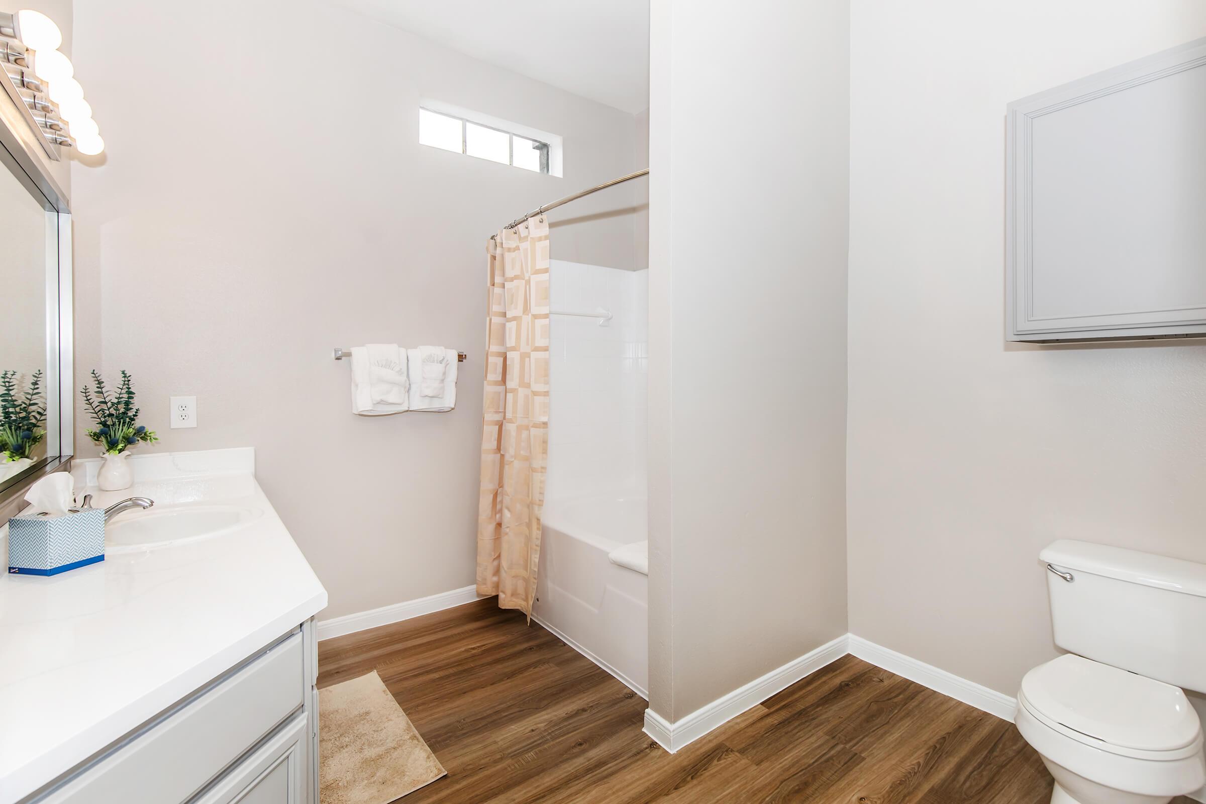 A modern bathroom featuring a shower with a beige curtain, a white bathtub, a white toilet, and a vanity with a large mirror. The walls are light-colored, and there's a small plant on the counter. Soft wood-like flooring completes the space, creating a clean and bright atmosphere.