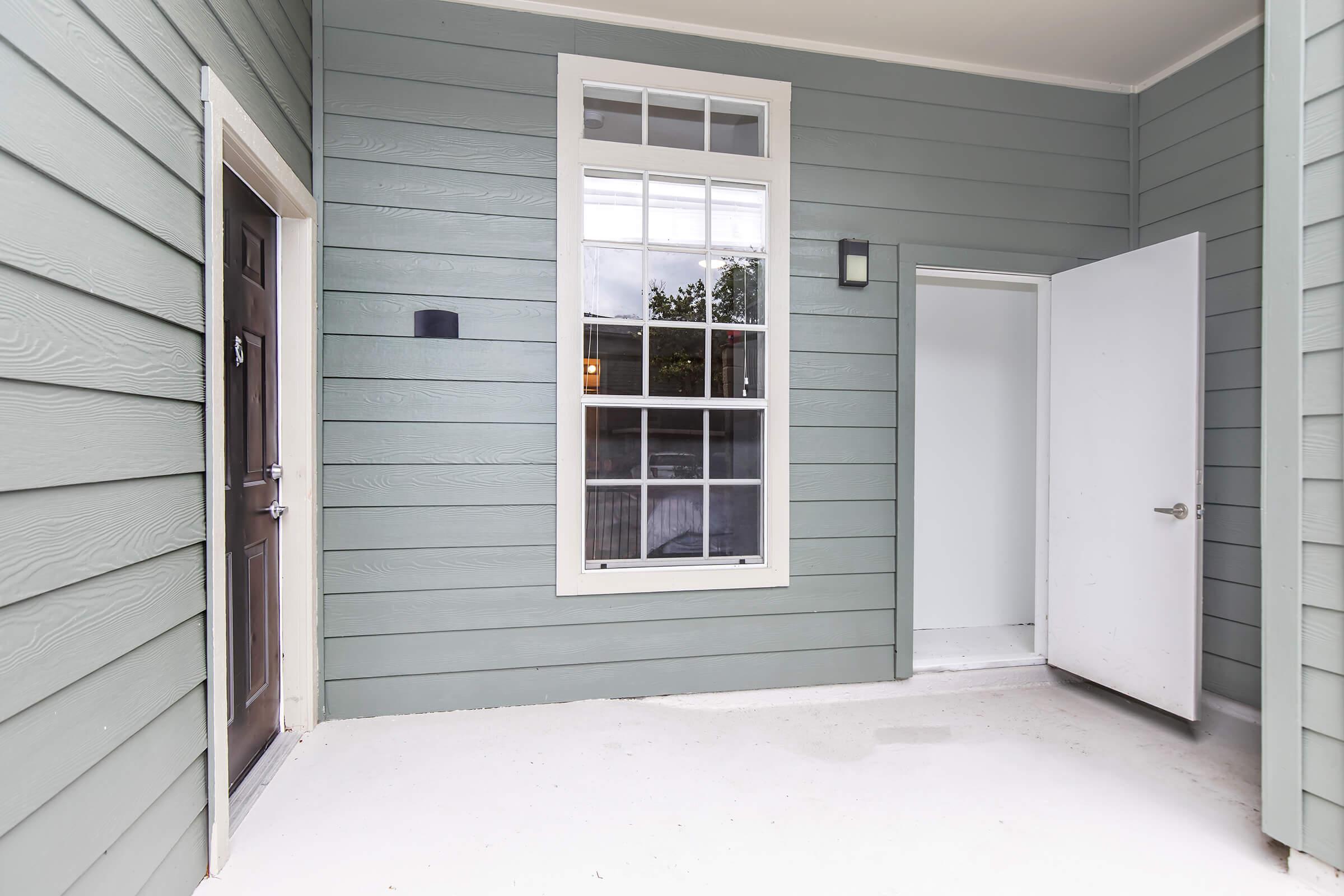 A corner of a building featuring light green siding, a window with multiple panes, and two doors: one on the left labeled with a number, and an open white door on the right leading to an interior space. The area is well-lit and has a clean, unfinished floor.