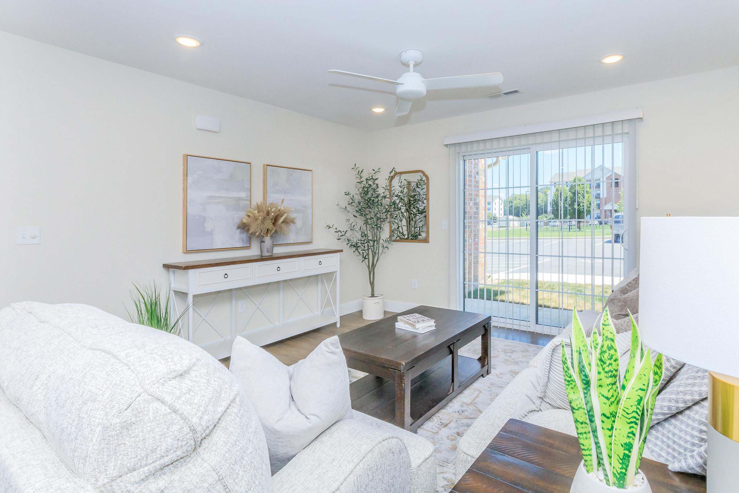 A bright and modern living room featuring a light gray couch, a wooden coffee table, and a decorative side table with a lamp and potted plant. There's a white console table with artwork above it and a large sliding glass door leading to an outdoor view. Natural light fills the space, creating a welcoming atmosphere.