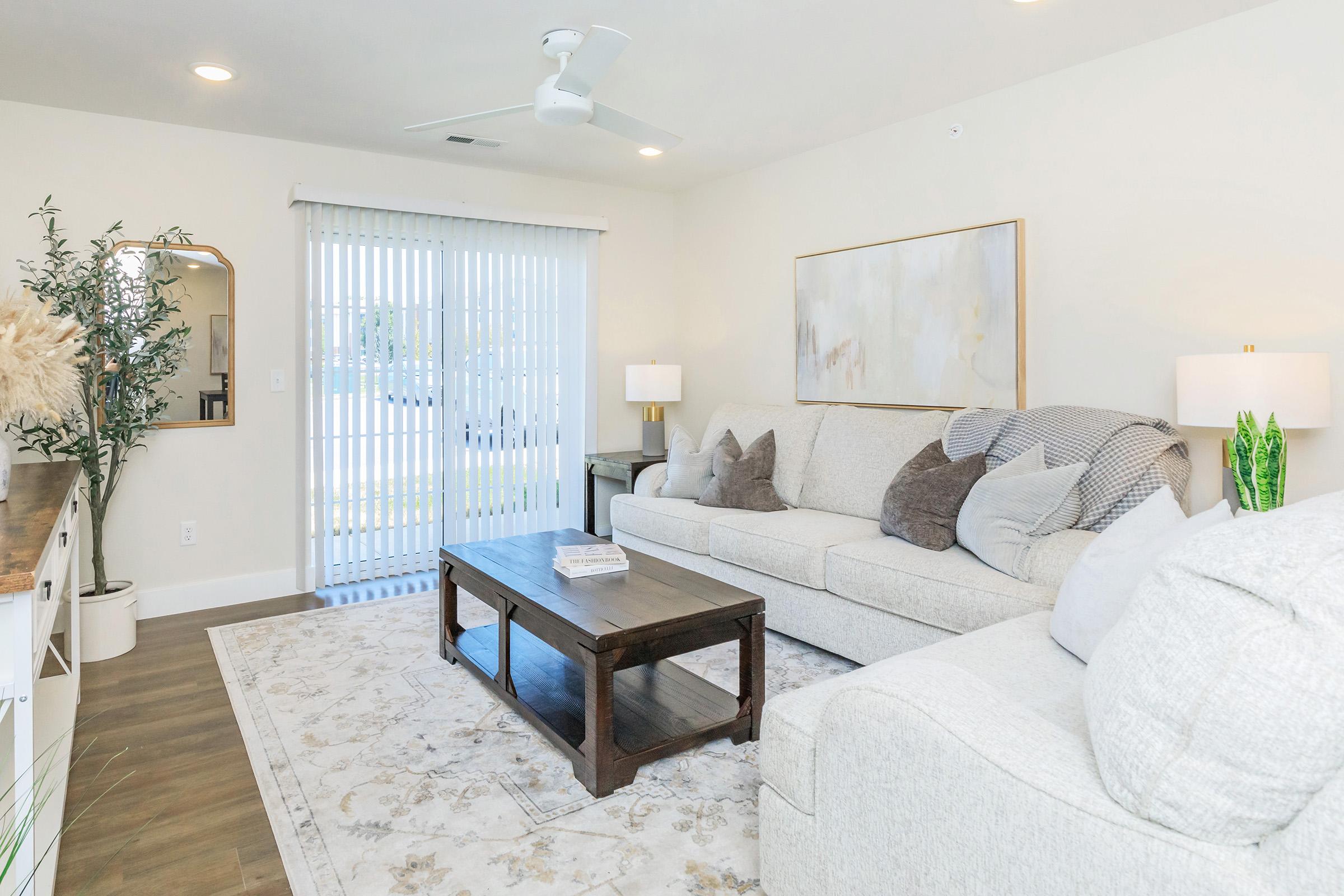 A cozy living room featuring a light-colored sectional sofa with decorative pillows, a wooden coffee table, and a large area rug. The space has soft lighting, a ceiling fan, and a window with vertical blinds, creating a warm and inviting atmosphere. A few plants and a decorative mirror add to the decor.