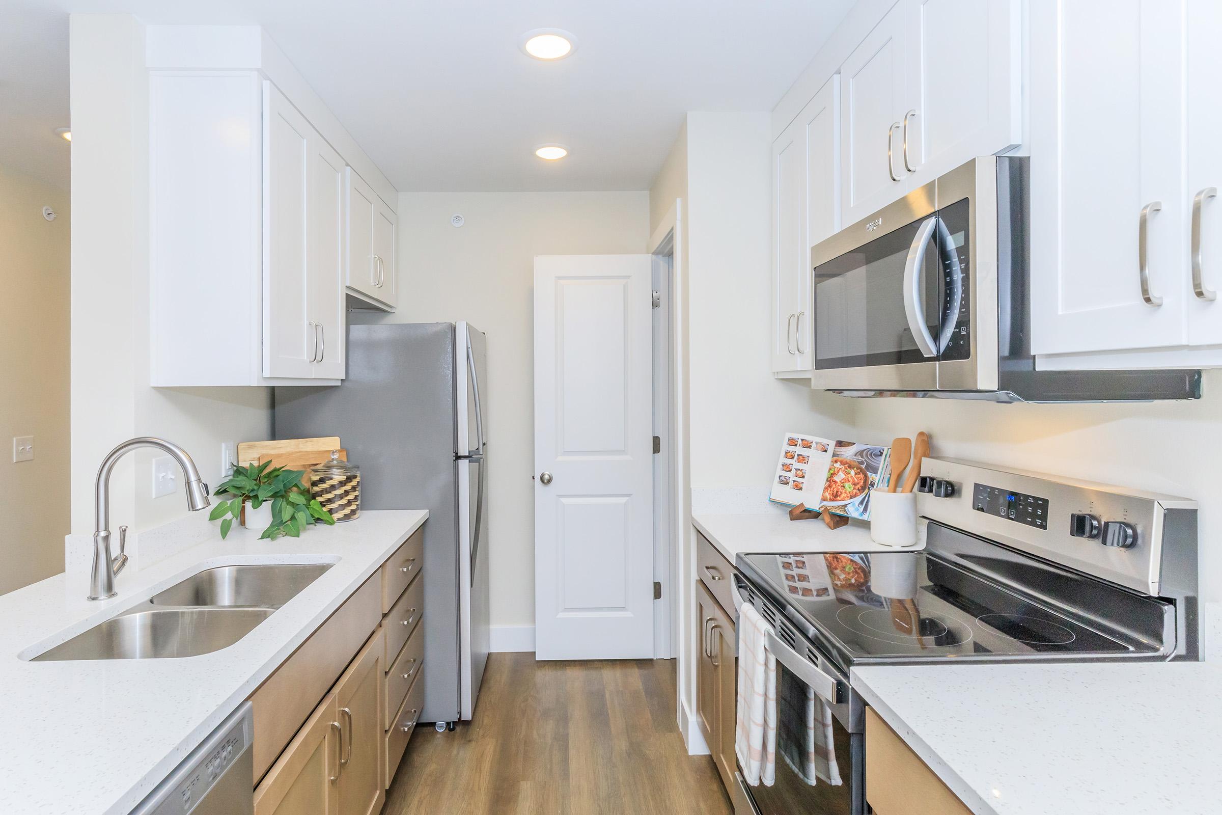 A modern kitchen featuring white cabinetry, stainless steel appliances including a refrigerator and oven, and a light-colored countertop. There’s a sink with a faucet and a small decorative plant. The room is well-lit with recessed lighting, showing a door leading to another area.