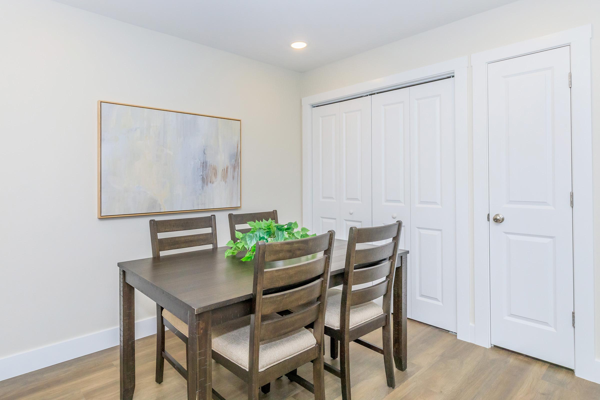A modern dining area featuring a wooden table with six chairs, adorned with a small green plant centerpiece. A large abstract painting hangs on the wall, and there are white double doors in the background. The space has neutral colors and a light, airy atmosphere.
