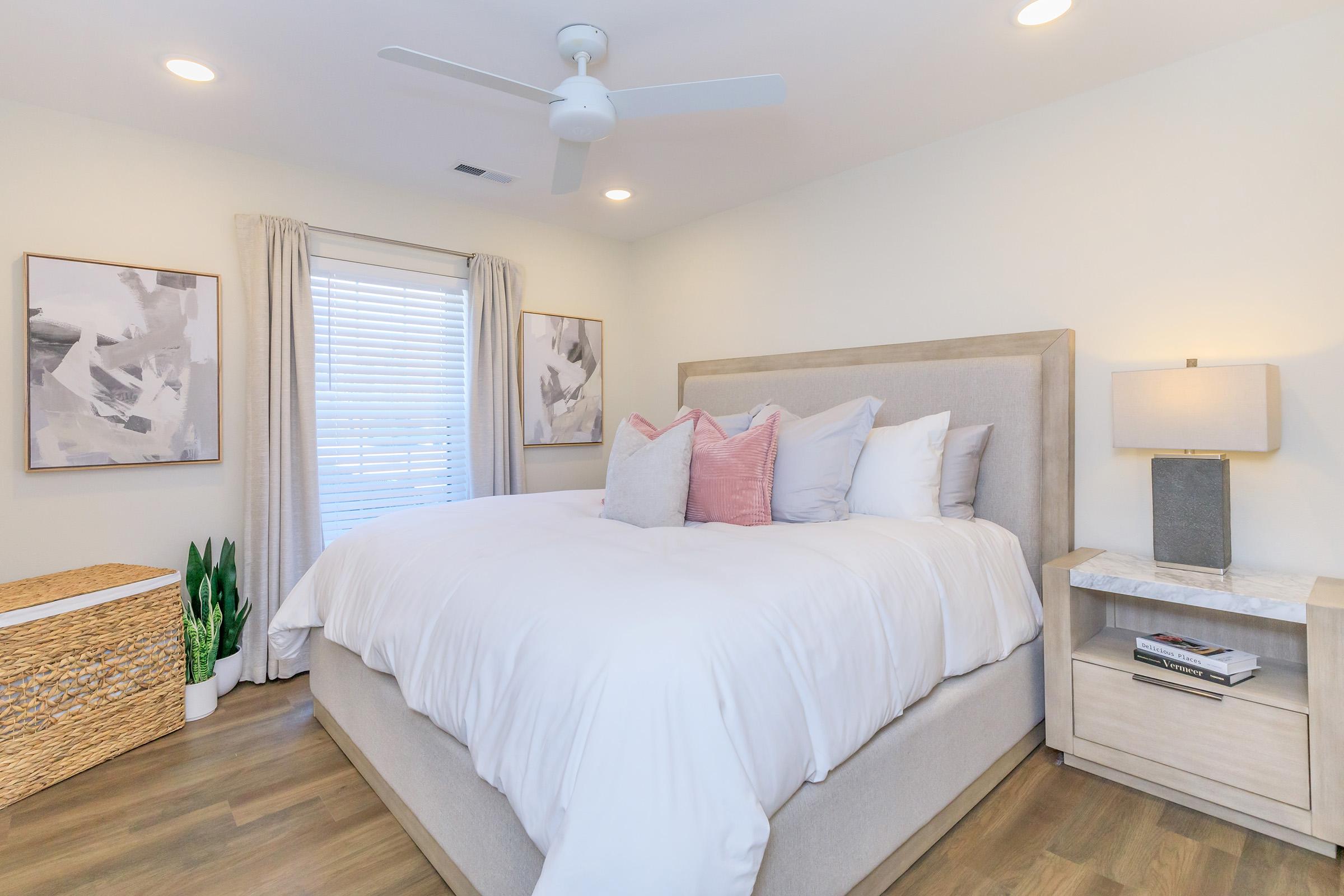 A cozy bedroom featuring a large bed with white bedding and decorative pillows in shades of pink and gray. There is a window with light curtains, a potted plant, and a bedside table with a lamp. Two abstract art pieces hang on the wall, and a woven storage basket is visible in the corner.