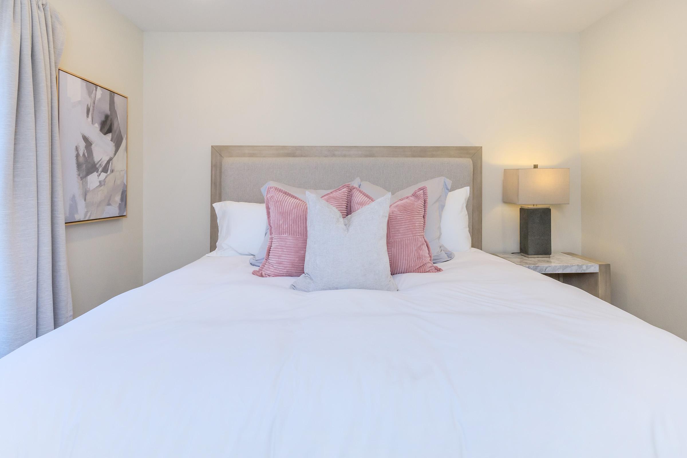 Cozy bedroom featuring a neatly made bed with white linens and decorative pillows in shades of pink and gray. A bedside table with a lamp is present beside the bed, and there's a piece of abstract wall art on the wall. Soft natural light illuminates the space, creating a warm and inviting atmosphere.