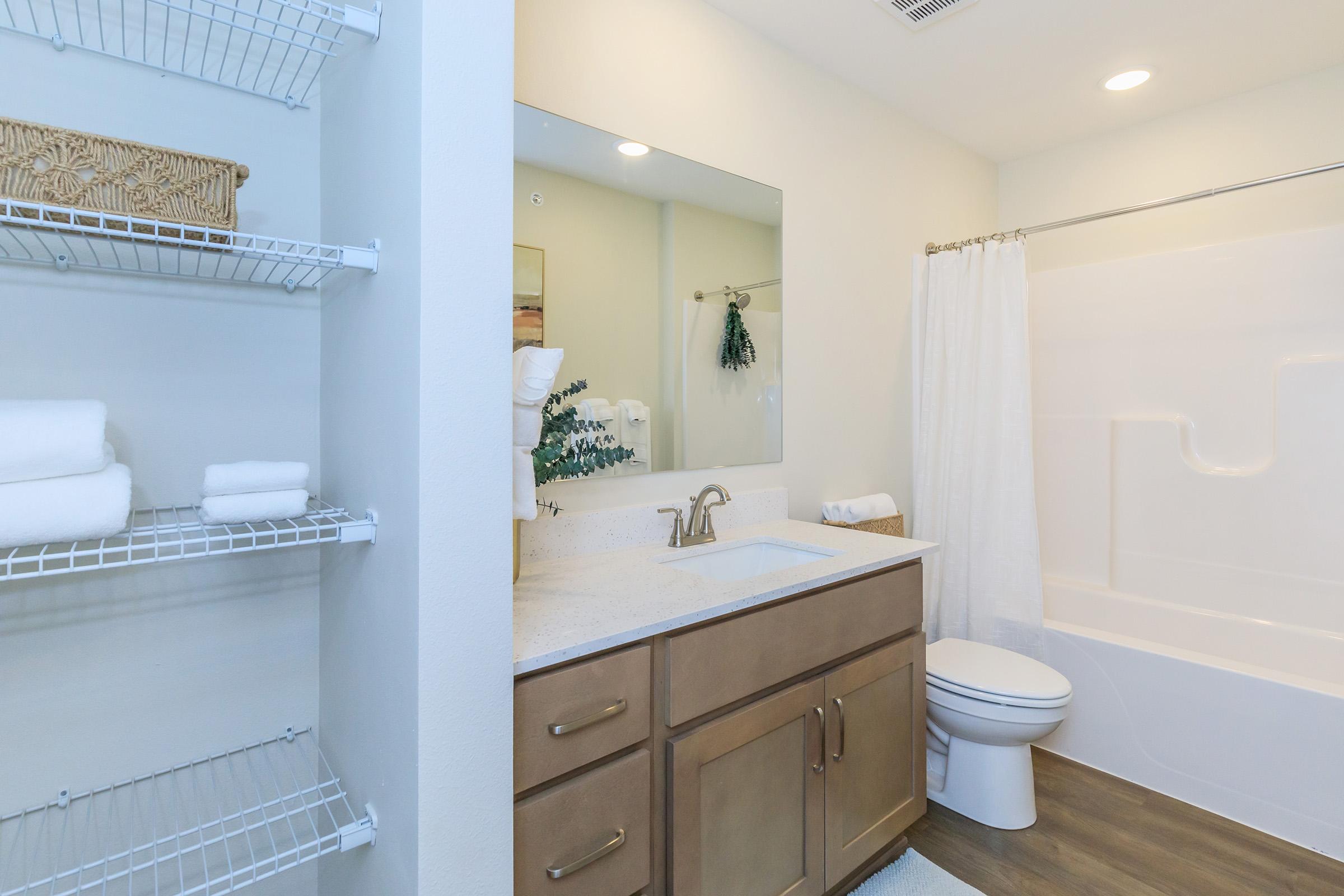 A clean and modern bathroom featuring a white bathtub with a shower curtain, a wooden vanity with a sink, and a large mirror above it. Shelves on the left hold neatly stacked white towels. The bathroom is well-lit, with neutral-colored walls and a simple decor.