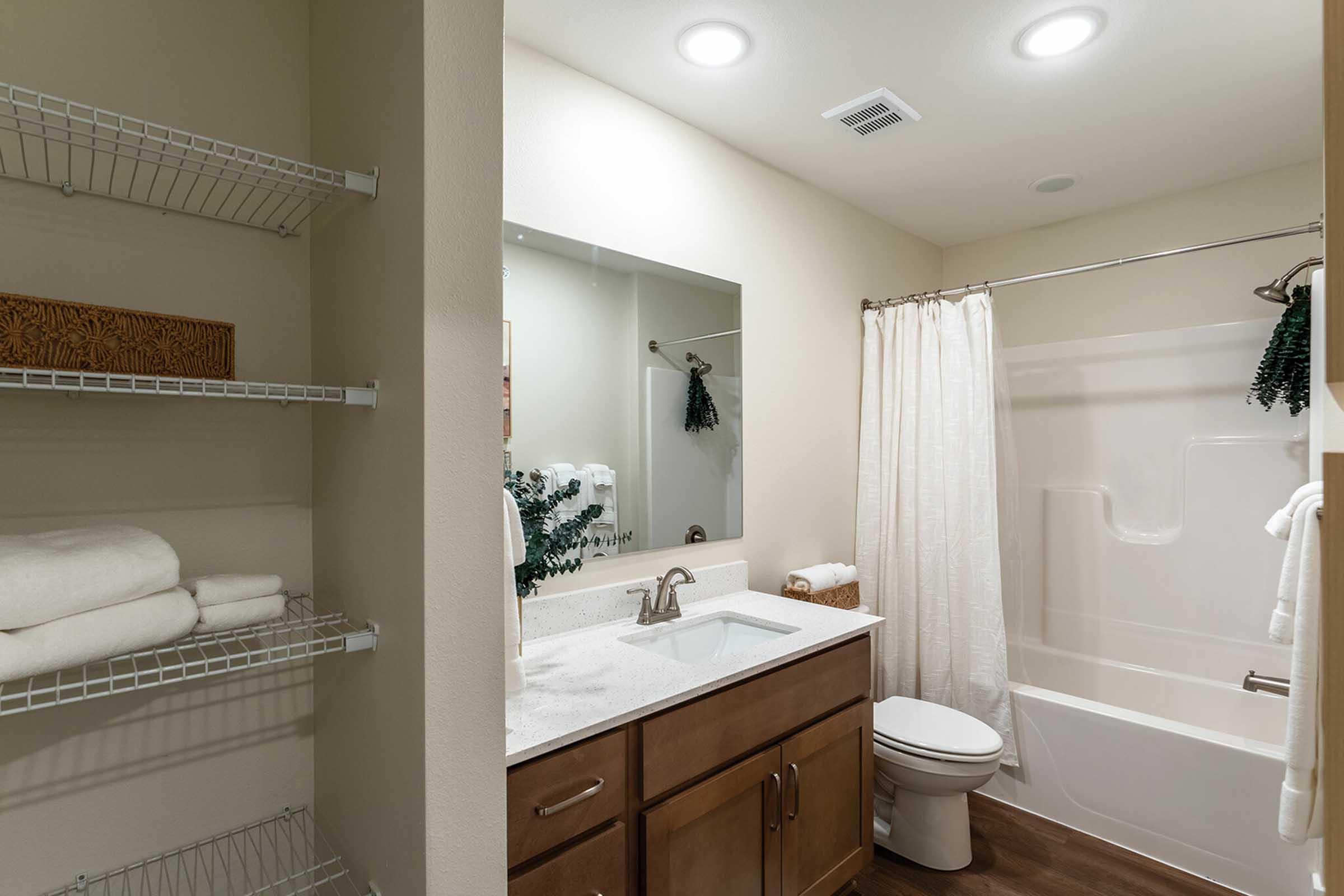 A modern bathroom featuring a white bathtub with a shower curtain, a sink with a dark wooden cabinet, and a mirror above it. To the side, there is a wire shelving unit with neatly stacked white towels. The walls are painted a soft beige, and soft lighting illuminates the space.
