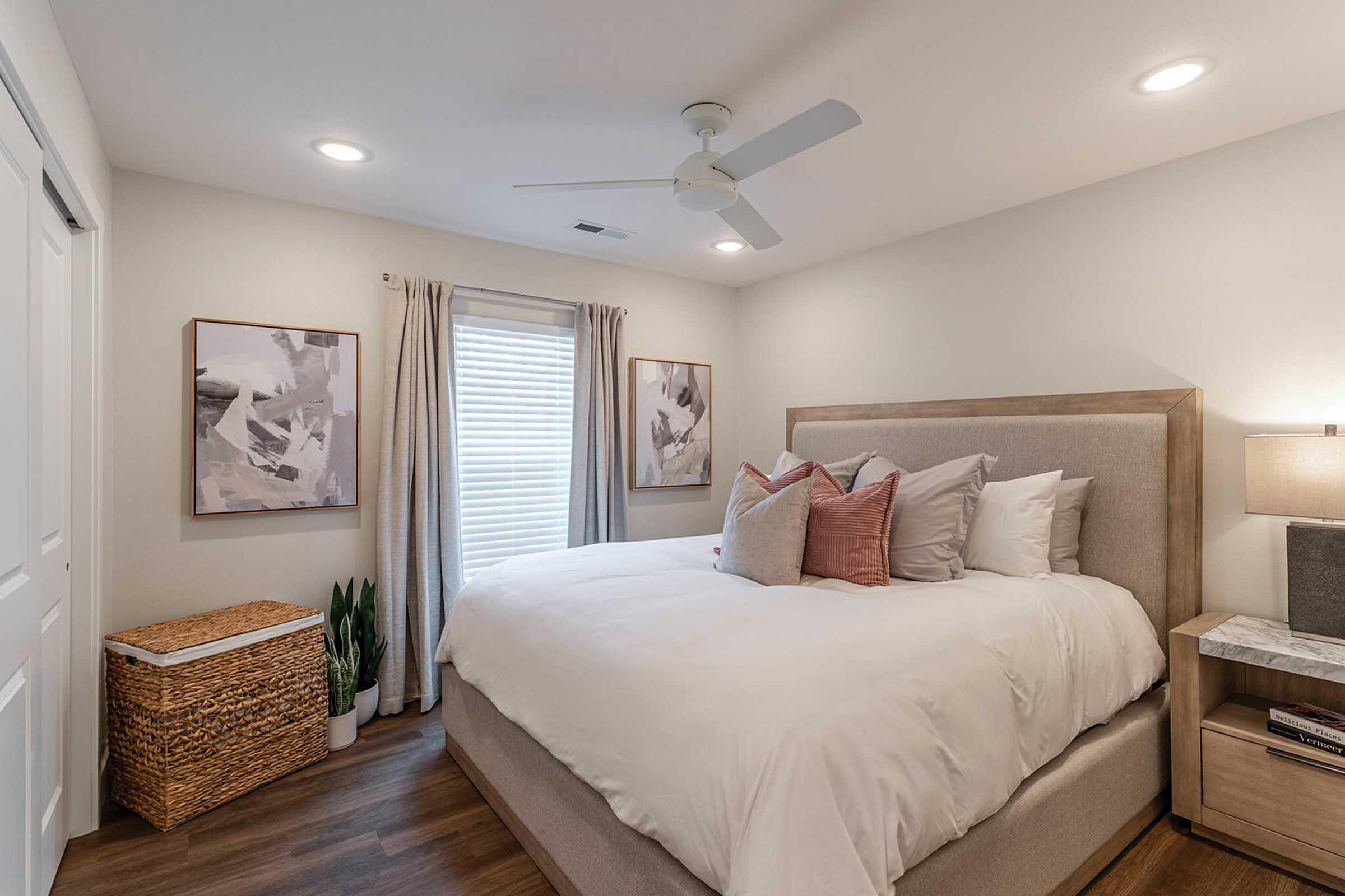 A cozy bedroom featuring a large bed with white bedding and decorative pillows. There are two abstract paintings on the walls, light-colored curtains, and a window with natural light. A woven basket and a small plant are in the corner, and a bedside table with a lamp is next to the bed. The flooring is wood.
