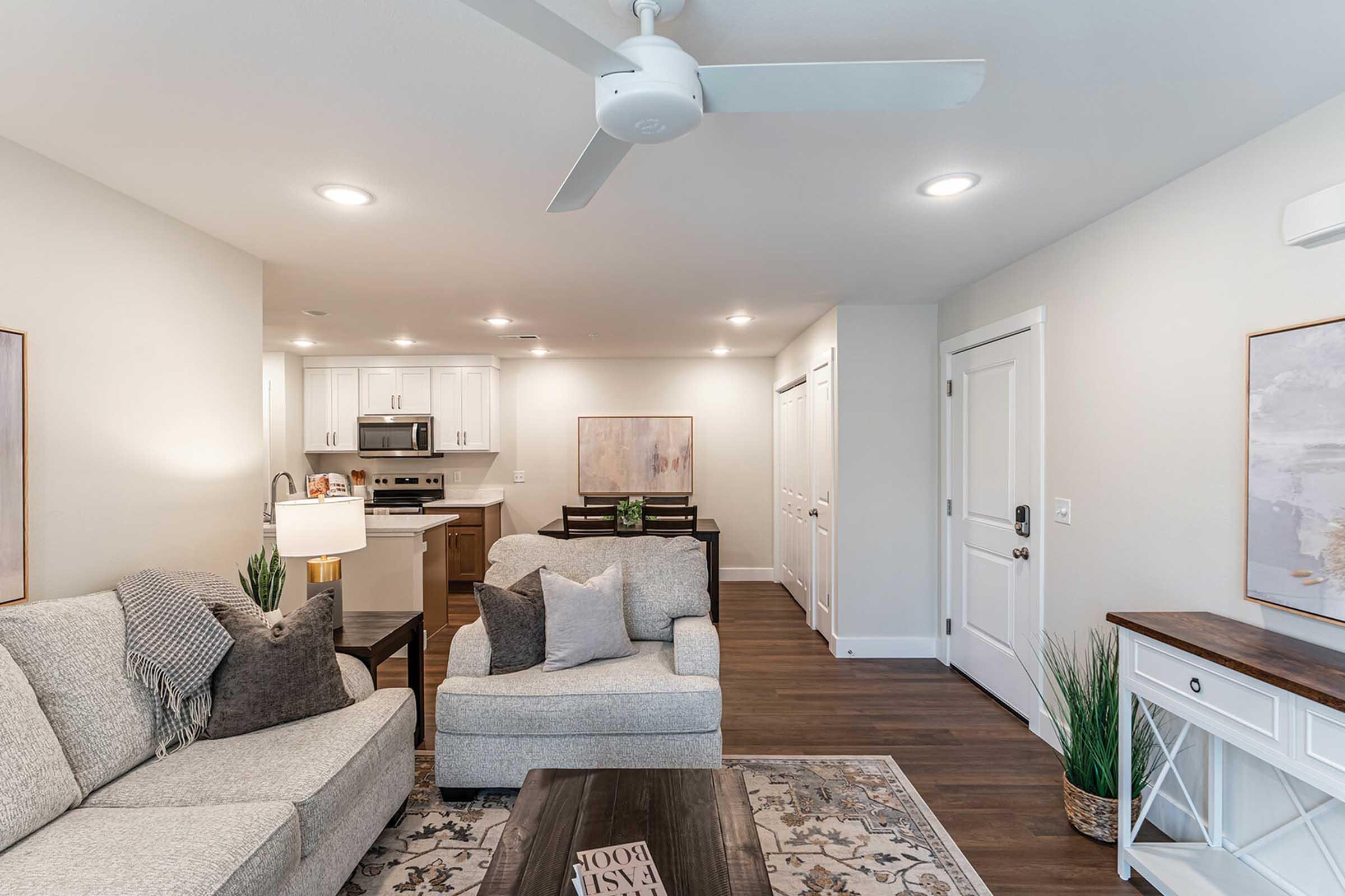 Cozy living room area with a light gray L-shaped sofa and decorative pillows, a wooden coffee table, and a floor lamp. The open space includes a kitchen with white cabinets and stainless steel appliances in the background, along with a dining area featuring a wooden table and a framed artwork on the wall.