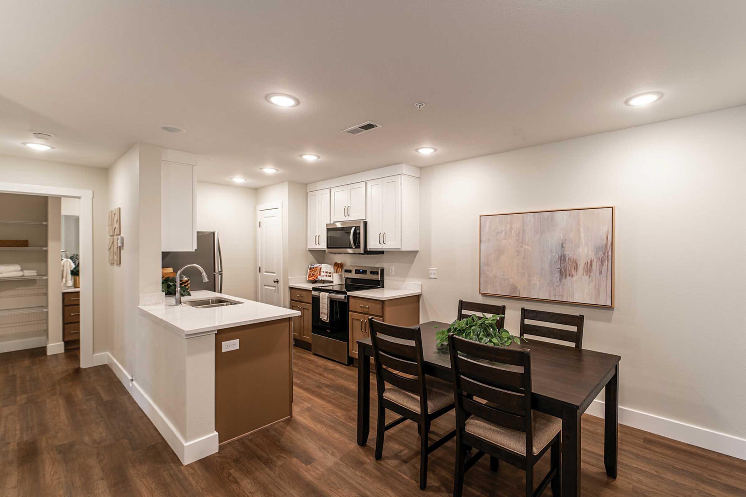 Modern kitchen and dining area featuring a white kitchen with stainless steel appliances, a wooden dining table with chairs, and warm lighting. The walls are painted in neutral tones, with decorative art hanging. A small pantry area is visible in the background. The flooring is a rich wood finish.