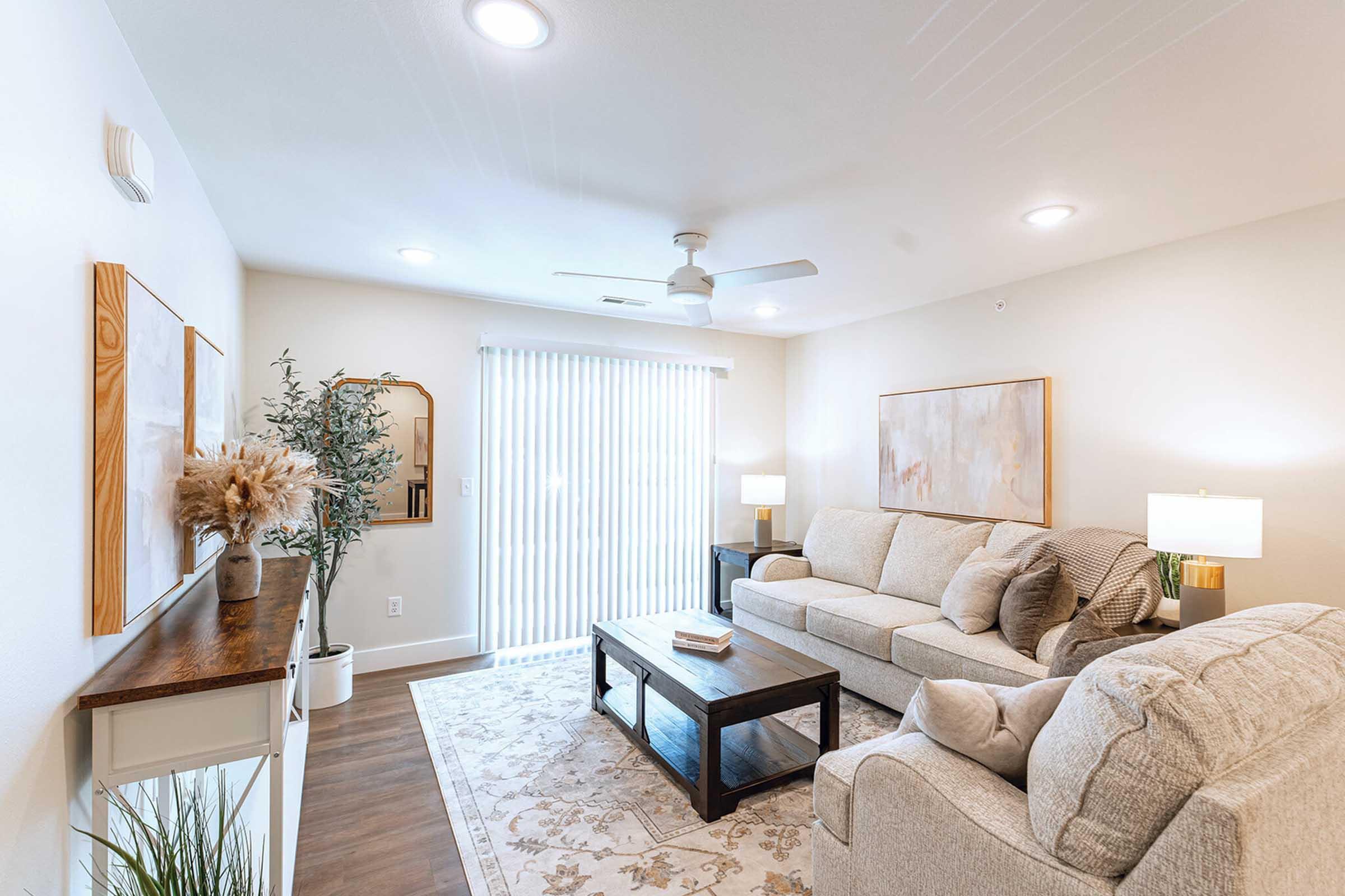 A cozy living room featuring a light-colored sofa with decorative cushions, a coffee table on a patterned rug, and a console table with decorative elements. There are two lamps providing soft lighting, a large mirror on the wall, and vertical blinds covering a window that lets in natural light.