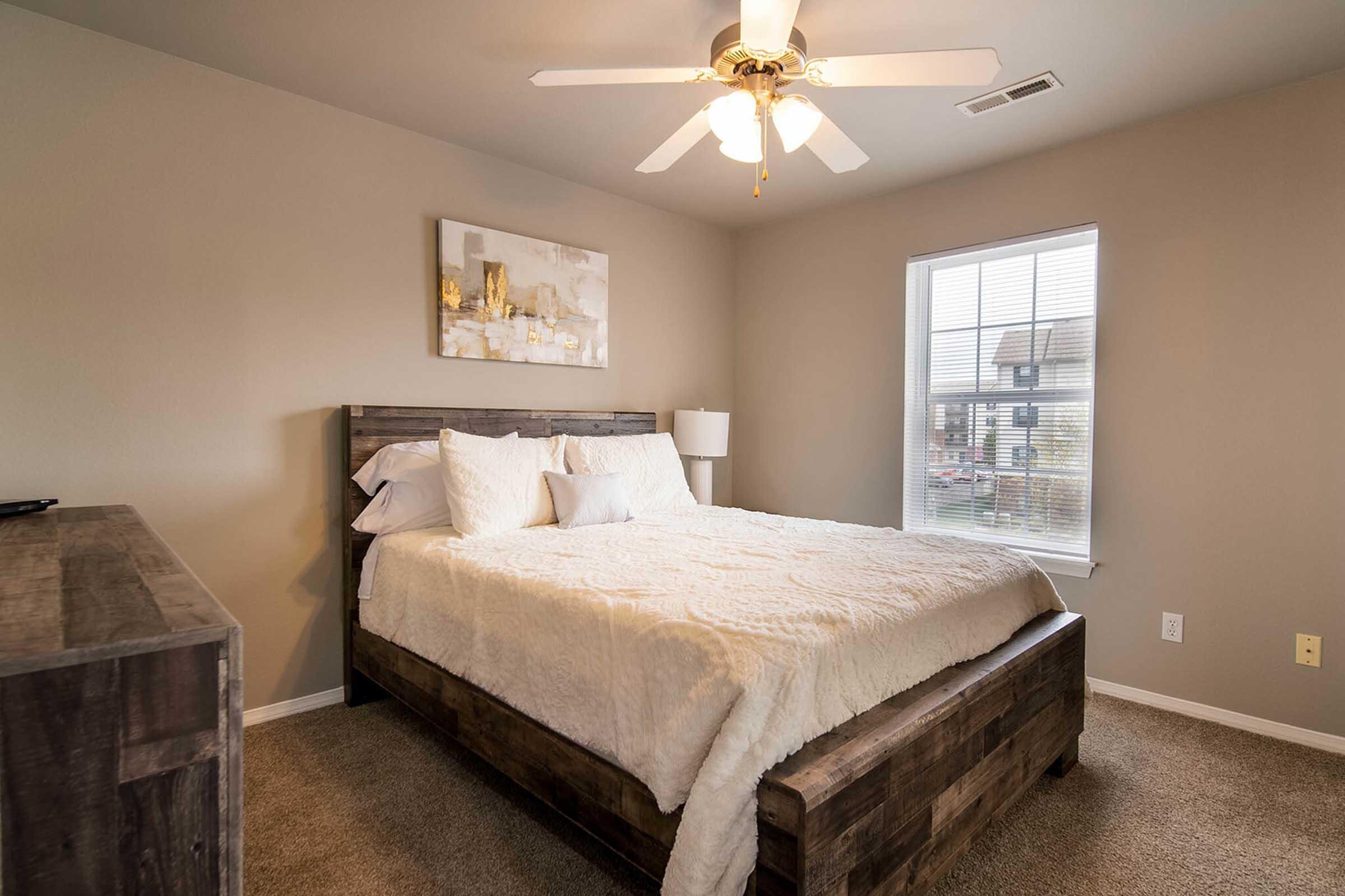 A cozy bedroom featuring a queen-sized bed with white bedding, a wooden headboard, and a matching nightstand. A ceiling fan provides airflow, and a large window allows natural light in. The walls are painted in a neutral tone, with a decorative artwork above the bed, creating a warm and inviting atmosphere.
