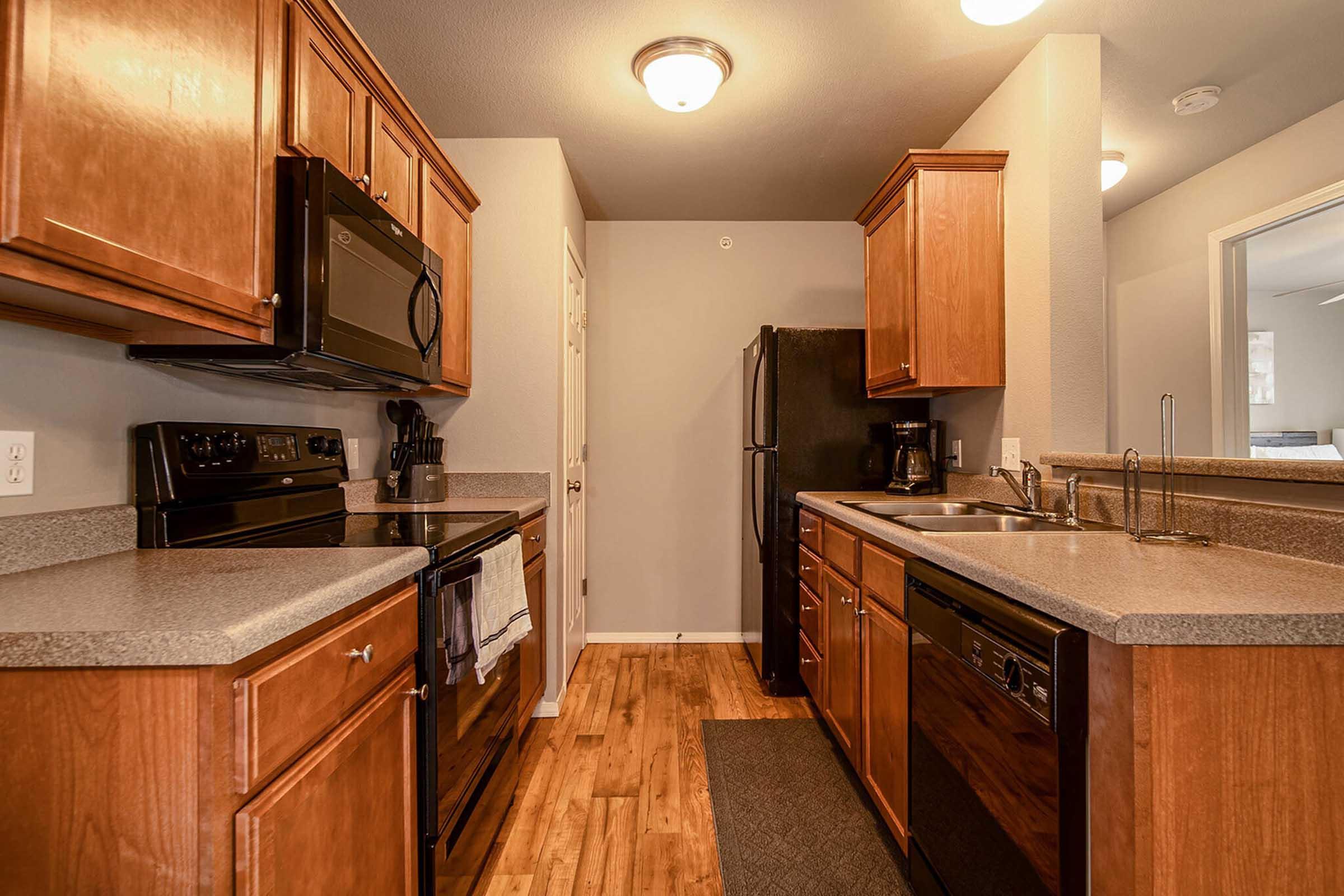 Modern kitchen with wooden cabinets, granite countertops, and stainless steel appliances. Features include a black microwave, oven, and refrigerator, along with a dishwasher. The flooring is wood laminate, and the walls are painted in neutral tones. The kitchen is well-lit with overhead lighting.