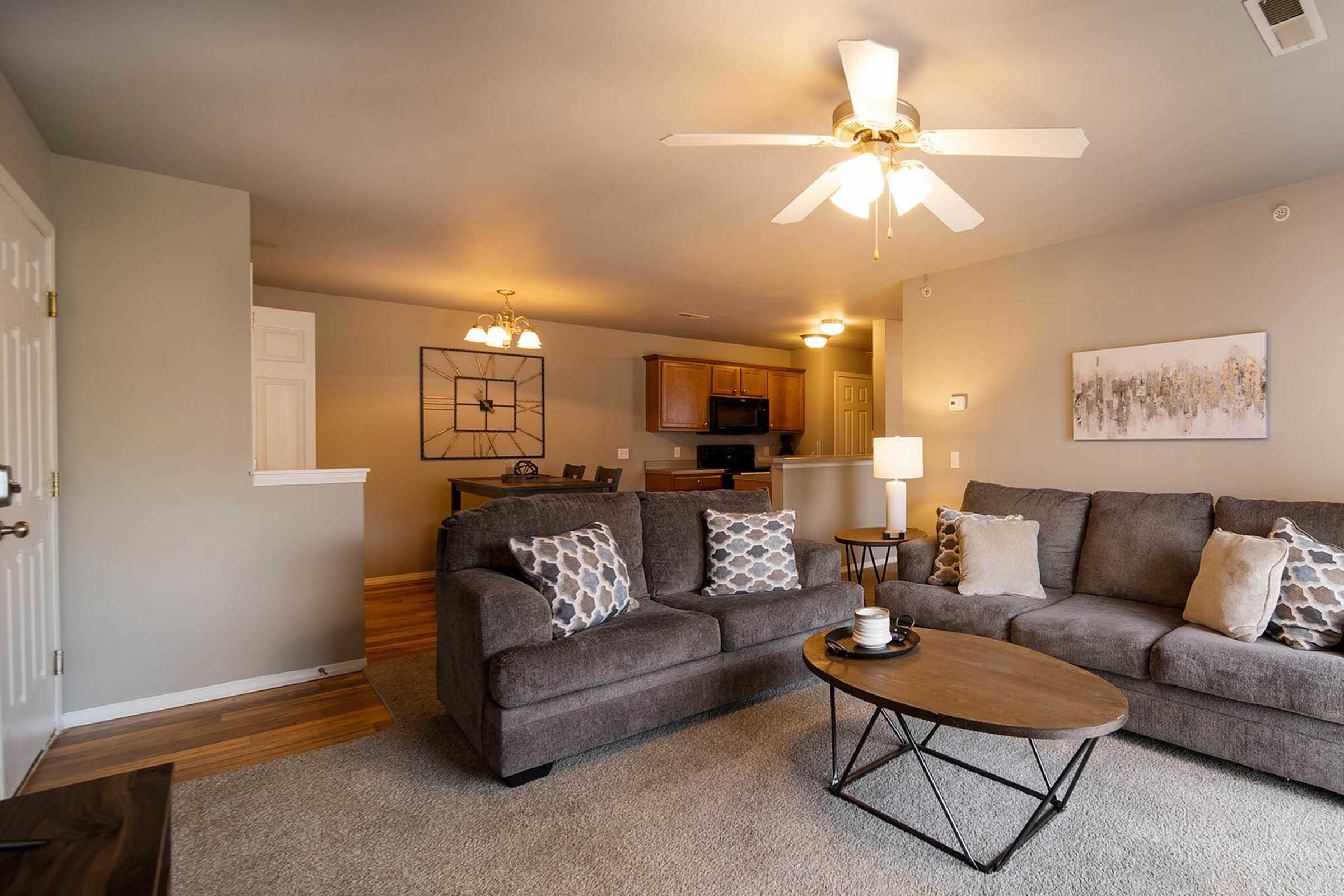Cozy living room featuring a gray sectional sofa with decorative pillows, a round coffee table, and a ceiling fan. The space is well-lit with warm lighting, and there’s an open view to a kitchen area in the background. Neutral wall colors and carpet enhance the inviting atmosphere.