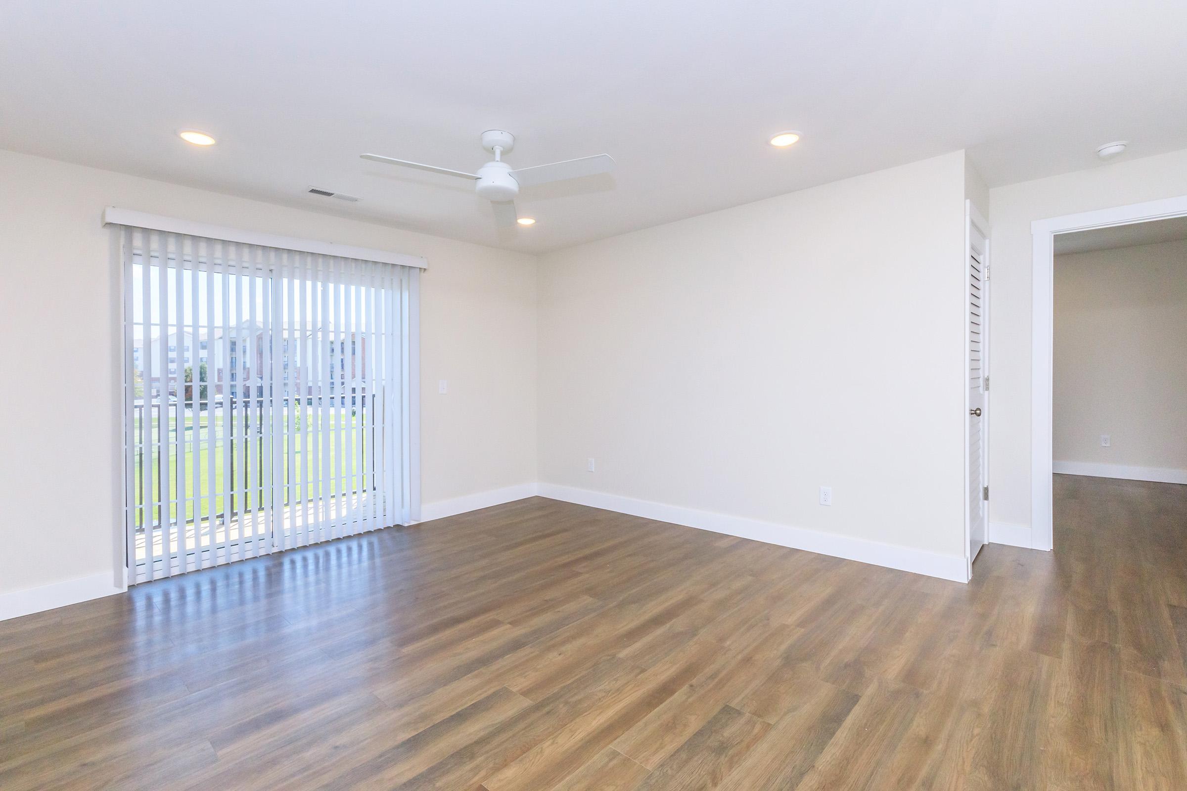 Bright and spacious room with light-colored walls and wood flooring. A ceiling fan is mounted on the ceiling, and there is a sliding glass door with vertical blinds leading to an outdoor area. The room is empty, providing a clean slate for decoration and furnishing.